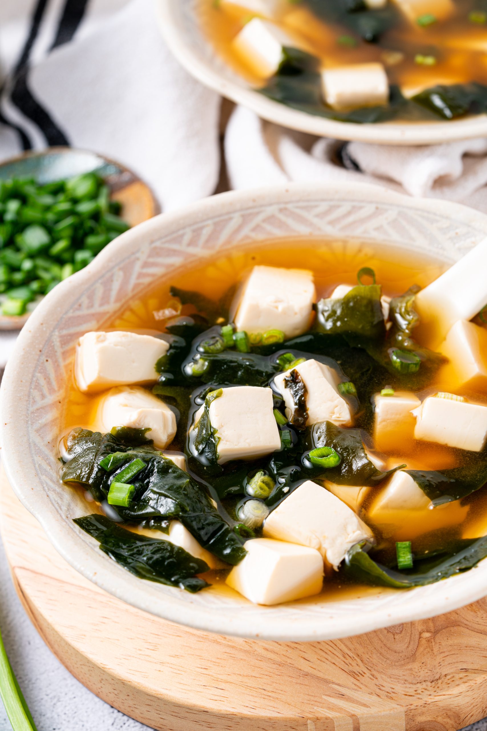 Side view of a bowl of miso soup with tofu and seaweed, garnished with green onions and served on a wooden board.