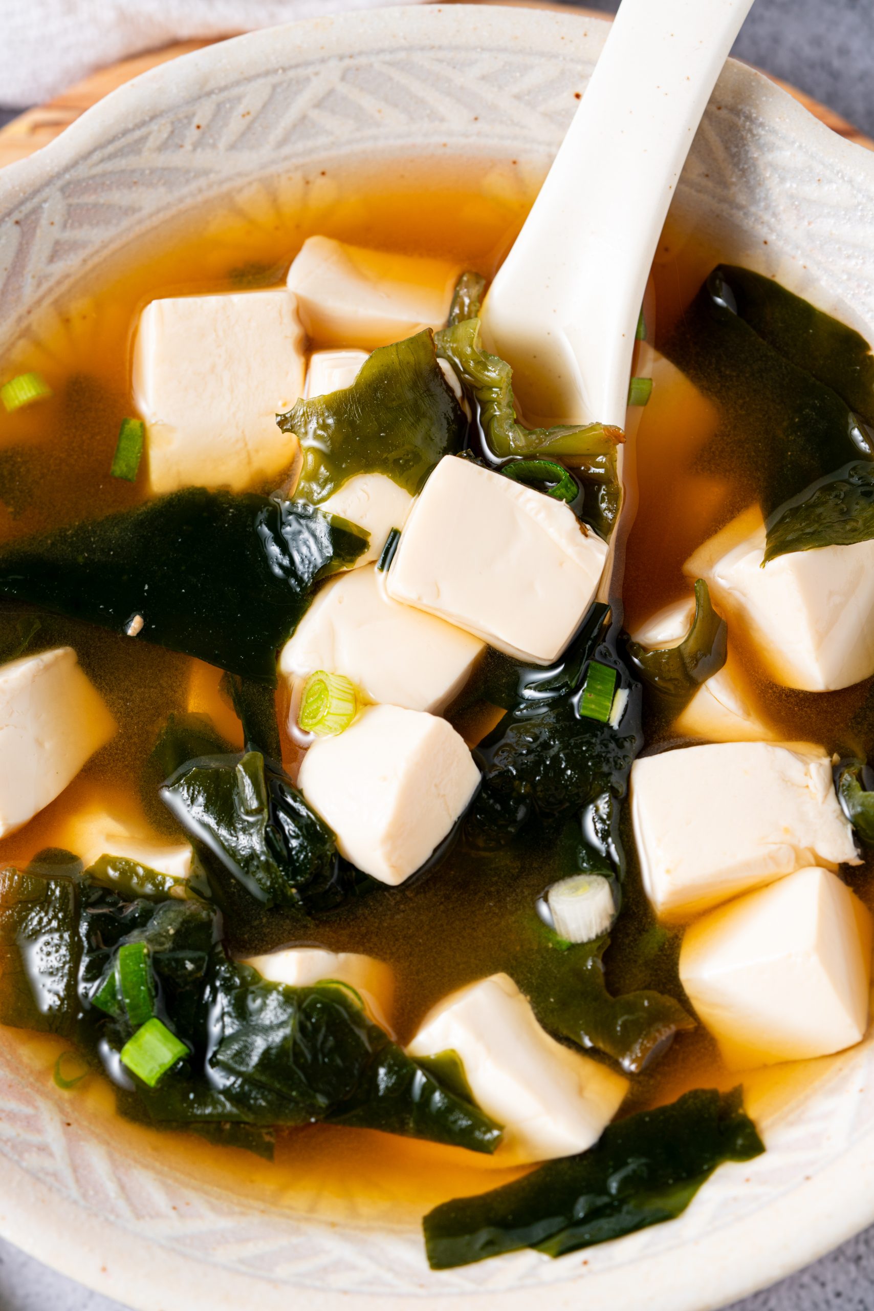 Spoon dipping into a bowl of Japanese miso soup, showing tofu cubes, wakame, and scallions in clear broth.