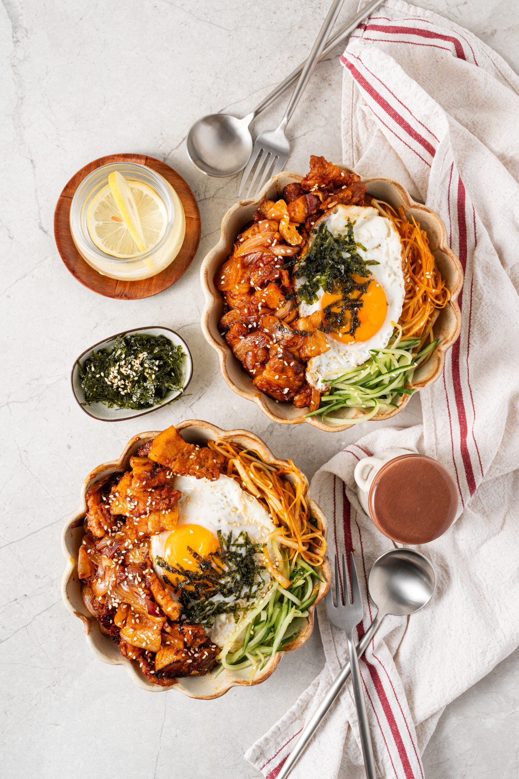Flat lay of two bowls of Korean pork stir fry with fried eggs and garnishes, surrounded by side dishes and utensils.