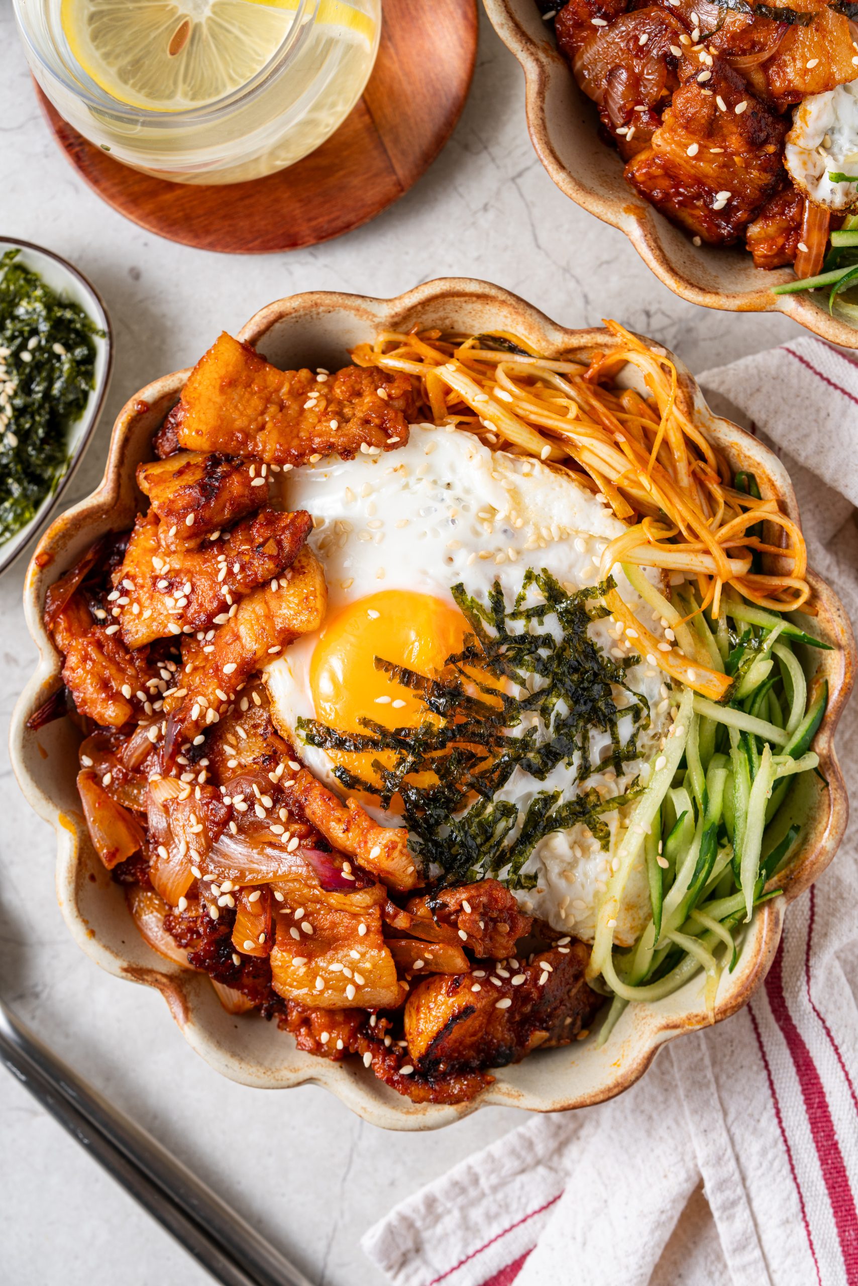 Korean pork stir fry served in a ceramic bowl with egg, cucumbers, and seasoned seaweed, styled with lemon water on the side.