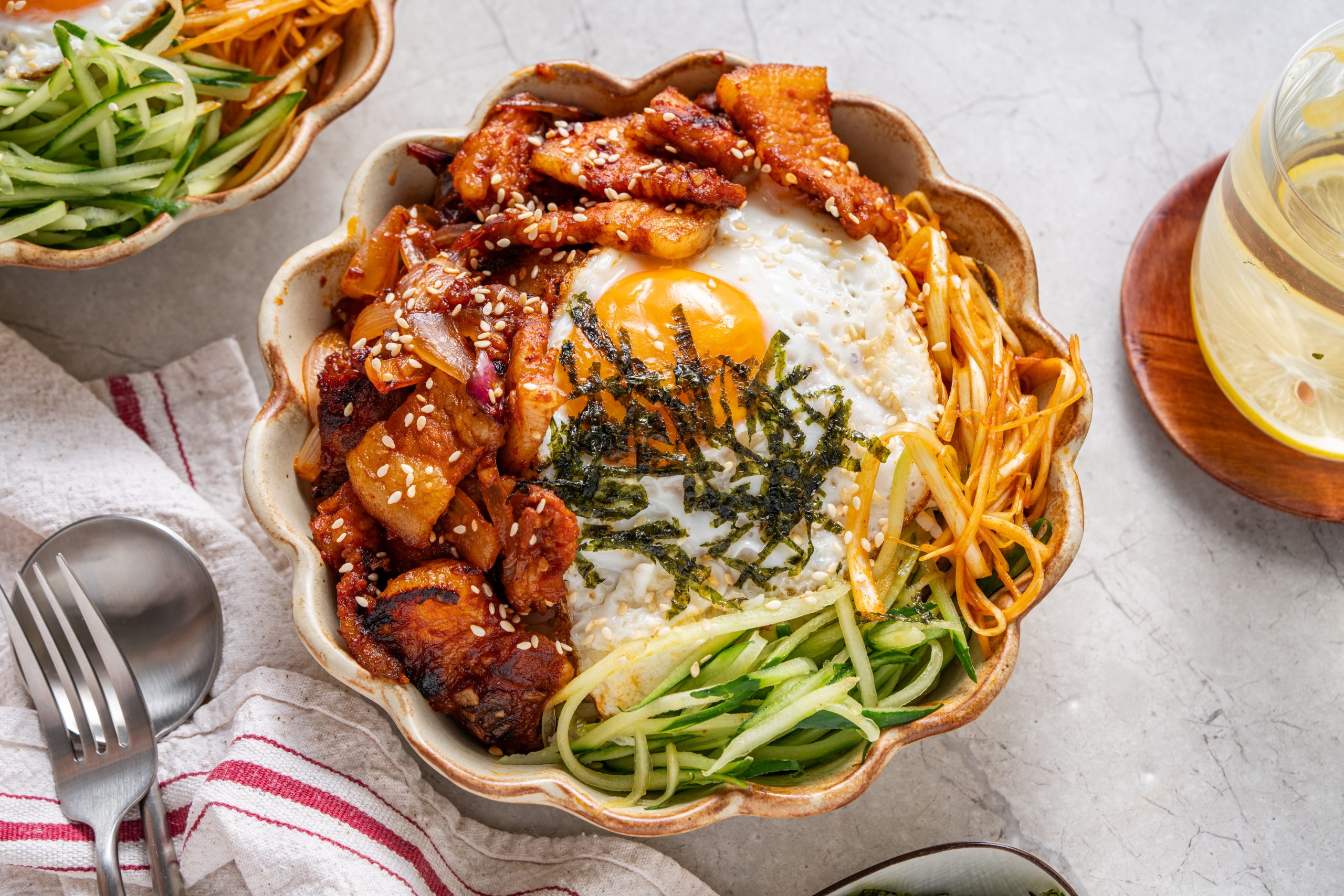 Overhead view of Korean pork stir fry bowl with fried egg, cucumber strips, and spicy noodles on a marble table.