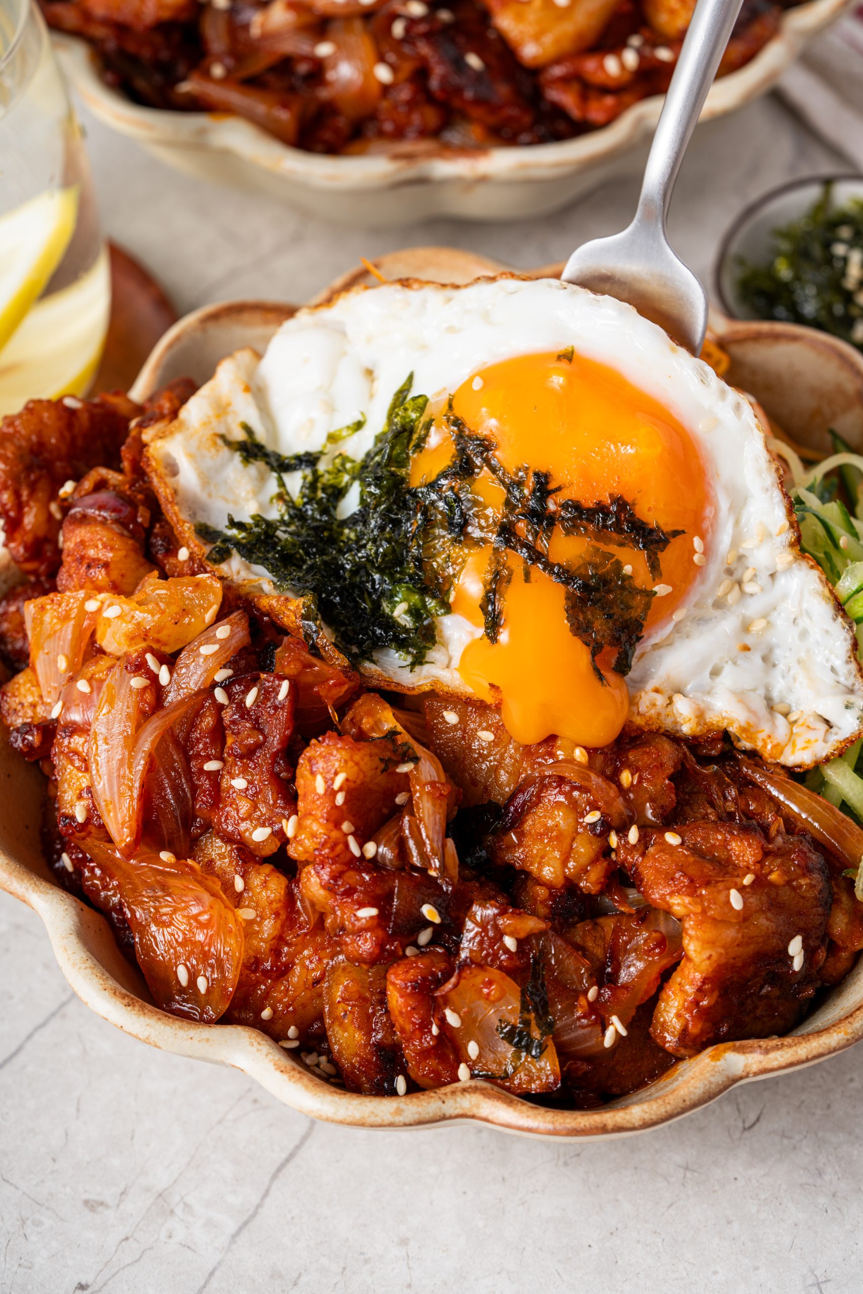 Korean pork stir fry bowl topped with a sunny-side-up egg, seaweed, and sesame seeds, served with sliced cucumbers.