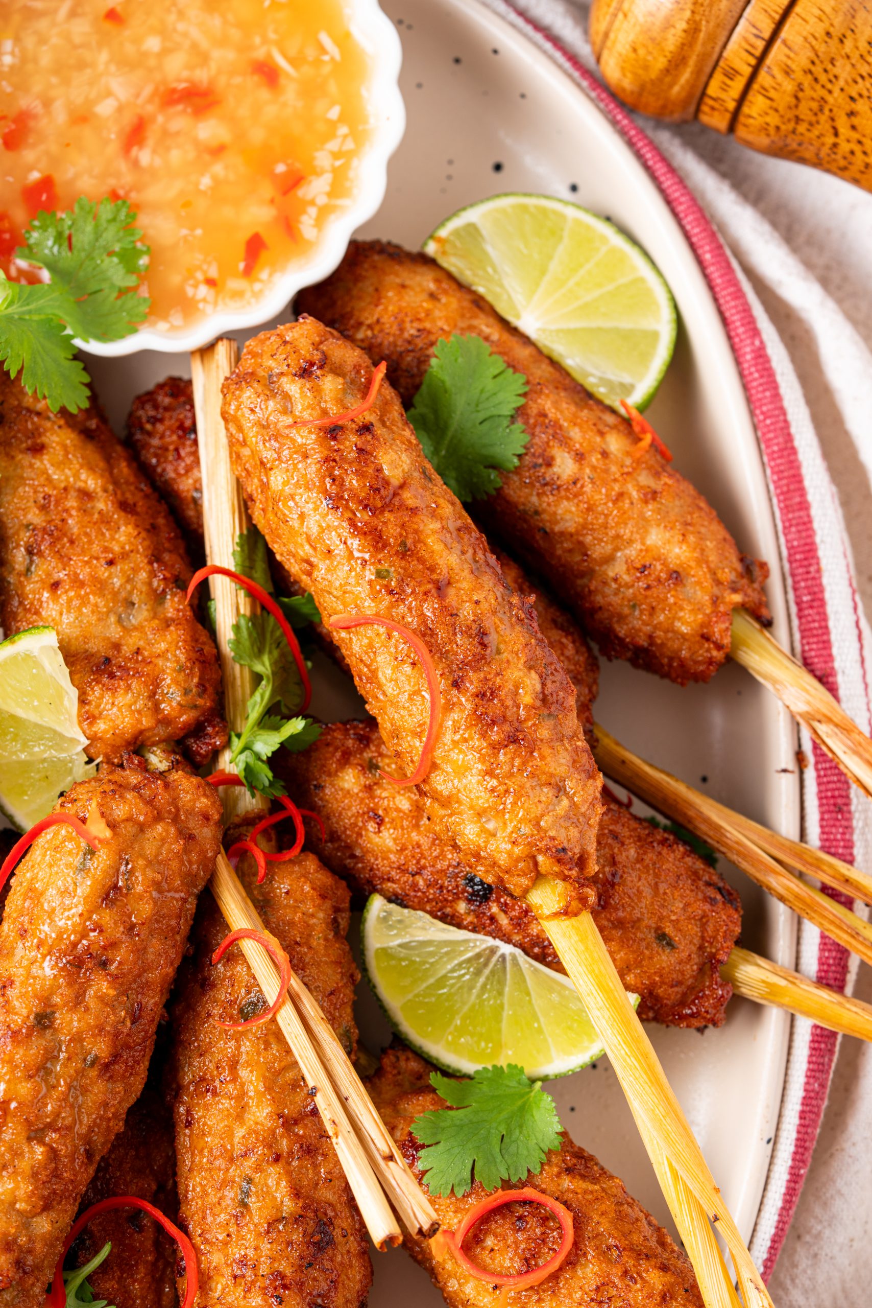A close-up shot of several lemongrass chicken skewers on a plate, showing their crispy browned exterior, garnished with lime wedges, cilantro, and sliced red chili near a bowl of sweet chili sauce.