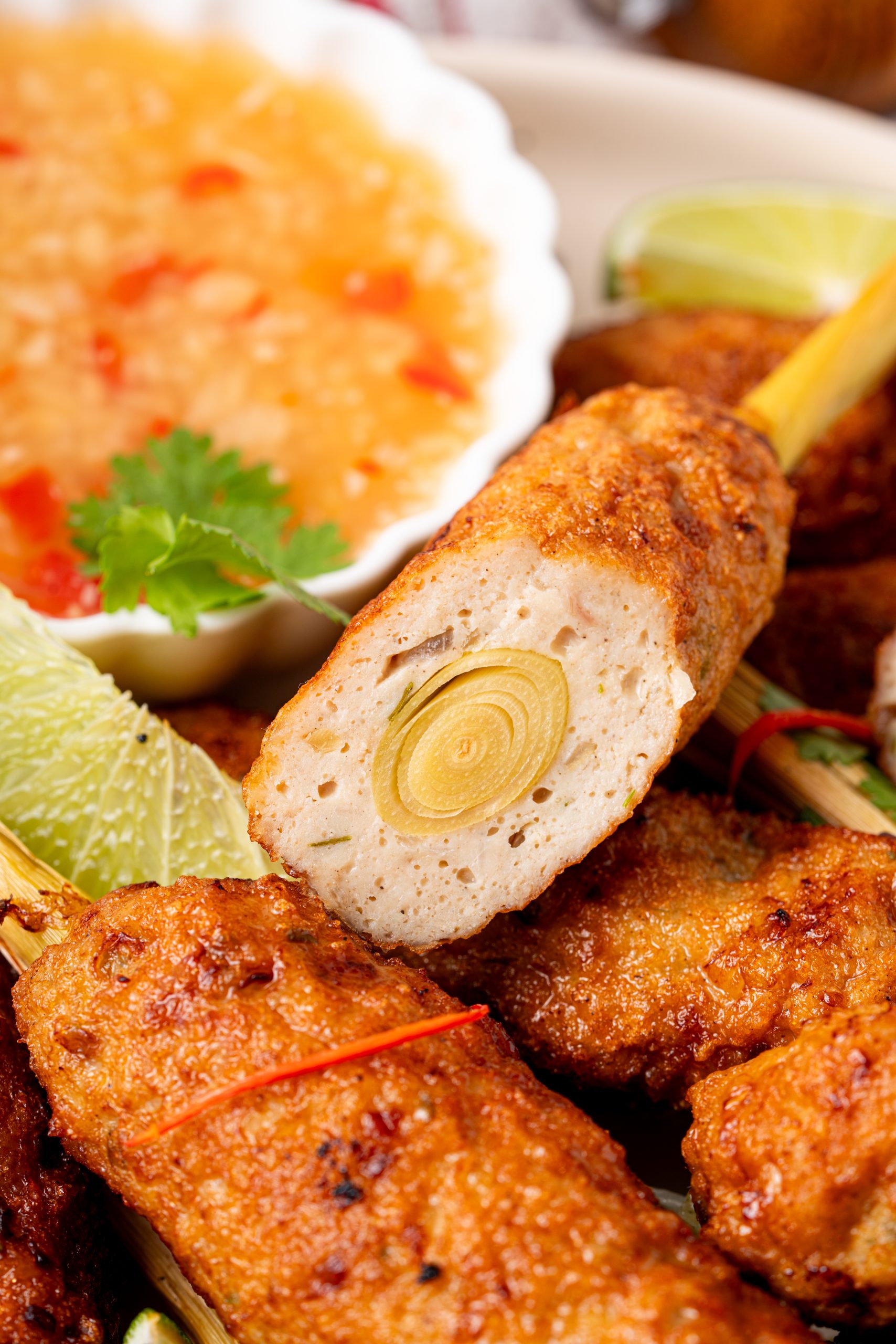 A cut-open lemongrass chicken skewer showing the inside texture and a visible lemongrass stalk at the center, placed beside a bowl of sweet chili dipping sauce with lime wedges and cilantro.