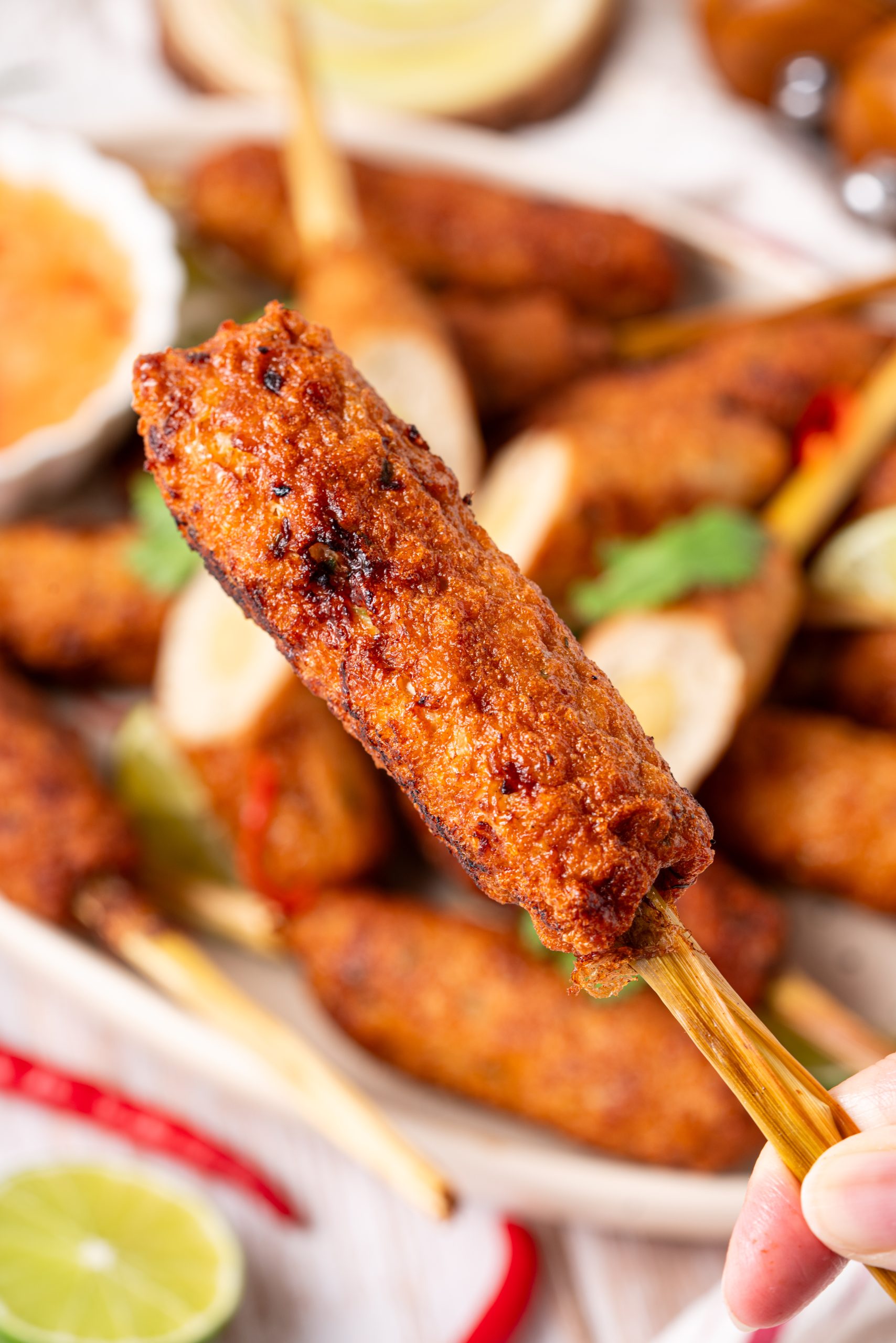 A hand holding up a crispy lemongrass chicken skewer, with a platter of skewers blurred in the background.