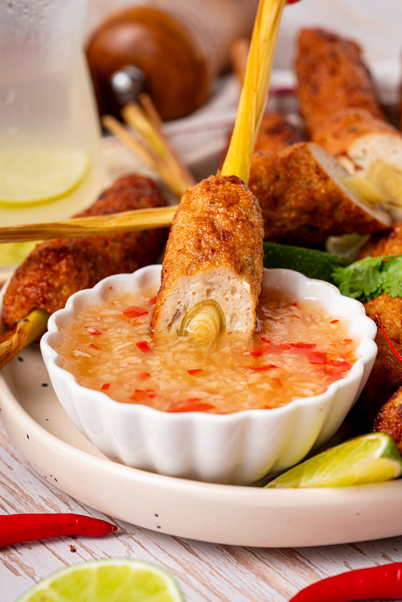 A lemongrass chicken skewer being dipped into a bowl of sweet chili sauce, with a plate of additional skewers and lime wedges in the background.