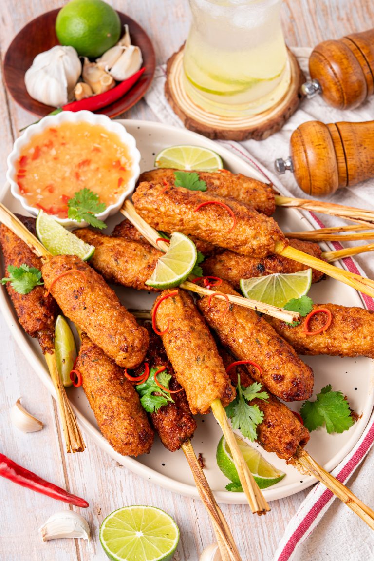 A platter of golden-brown lemongrass chicken skewers arranged on a large plate, garnished with lime wedges, sliced red chili, and fresh cilantro, accompanied by a small bowl of sweet chili dipping sauce and surrounded by garlic, chili, and a refreshing drink.