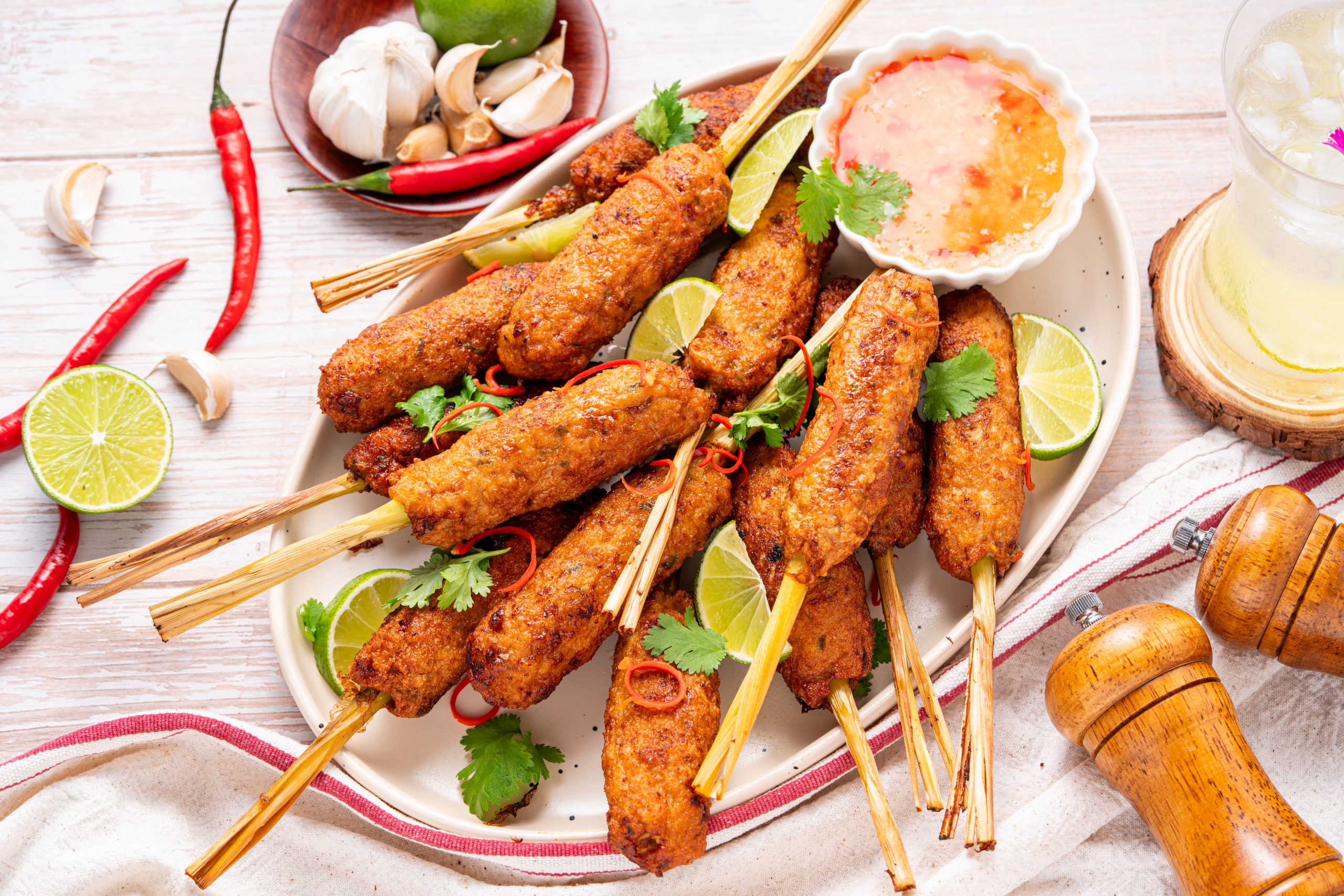 Top-view of a large plate filled with crispy lemongrass chicken skewers, garnished with lime slices, cilantro, and red chili strips, with fresh garlic, chili peppers, lime, and a drink placed around the plate.