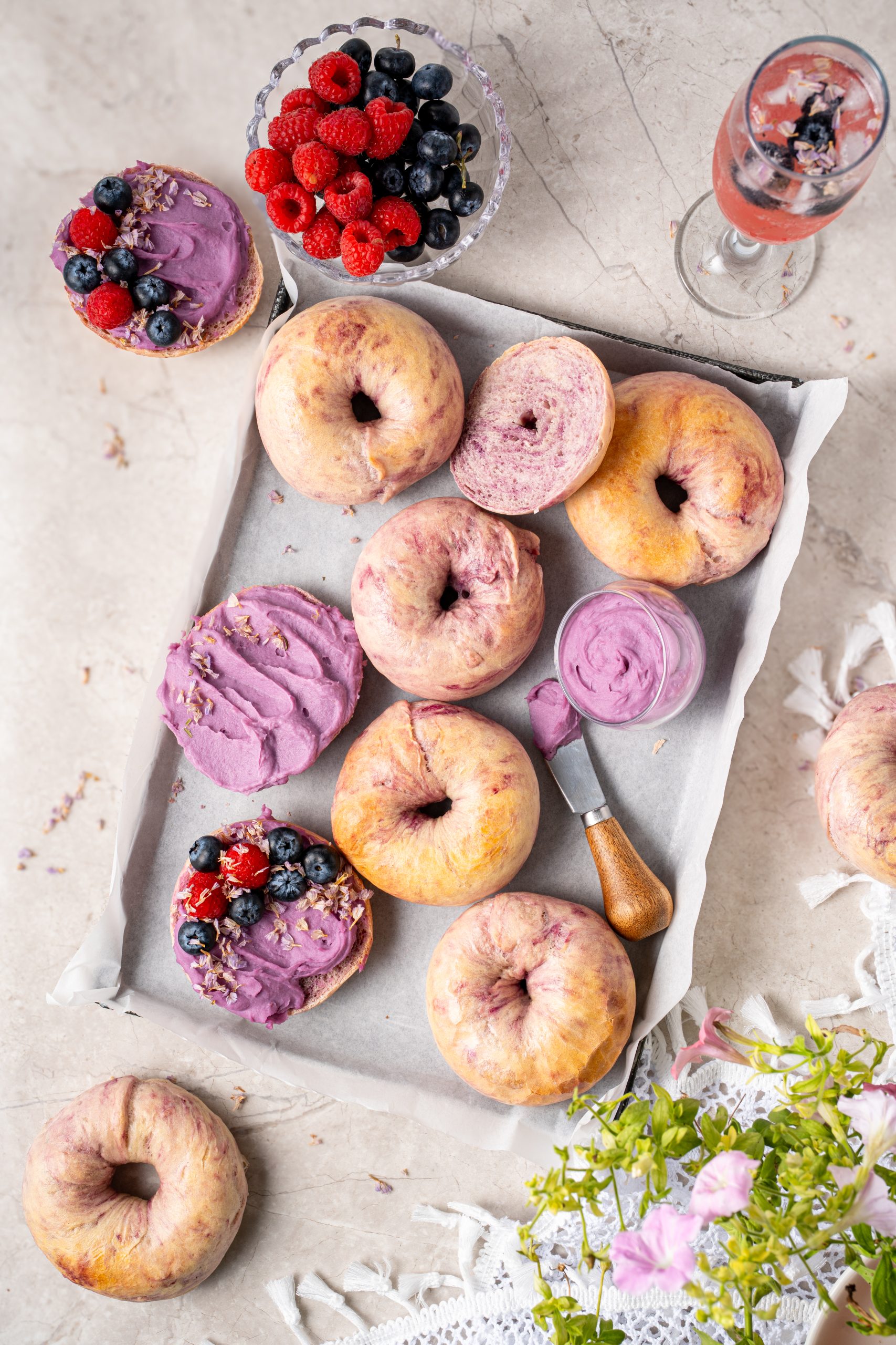 Overhead view of Purple Sweet Potato Bagels with creamy purple spread, raspberries, blueberries, and a glass of berry drink.