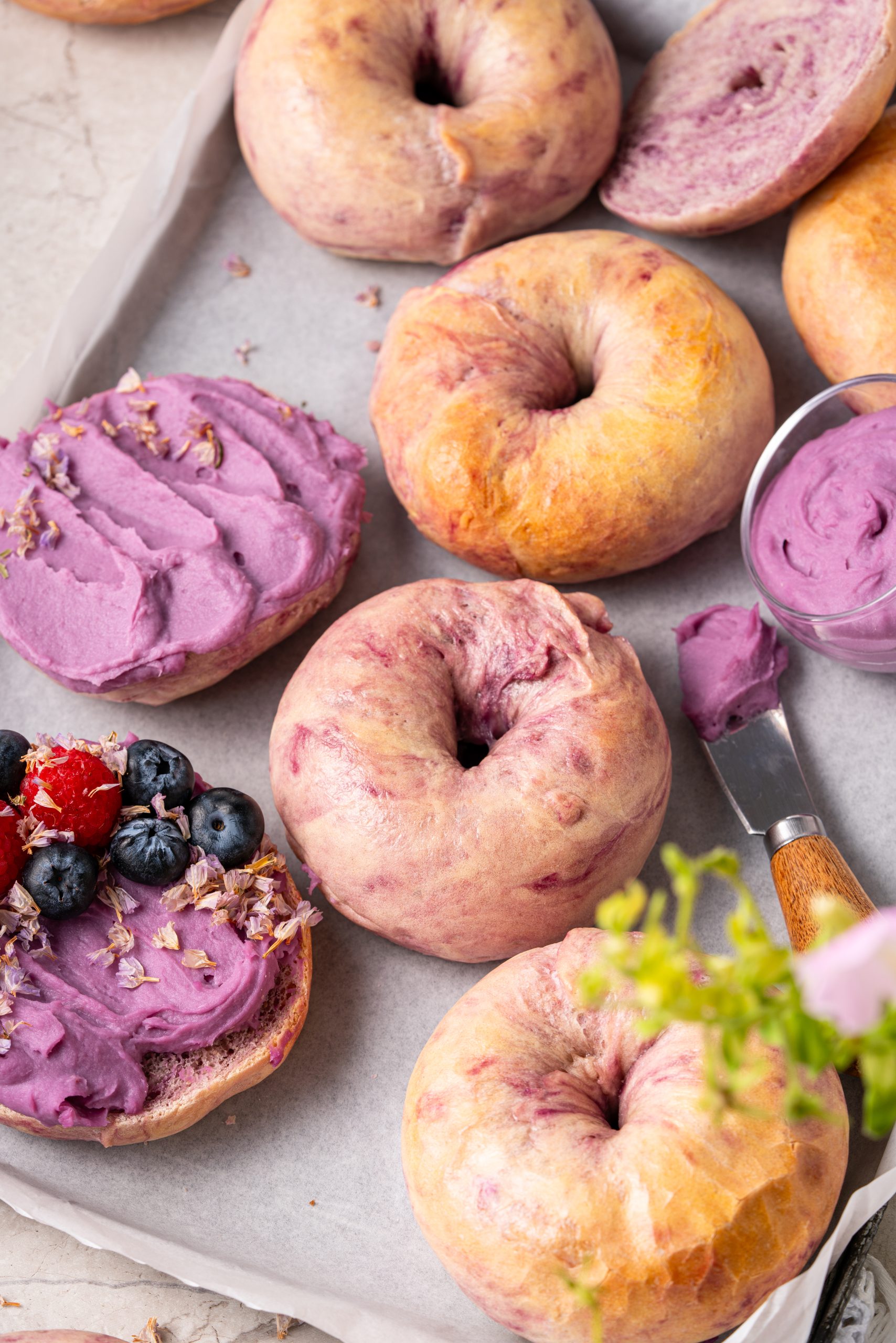 Overhead view of purple sweet potato bagels on parchment paper, with whole and sliced bagels showing a soft purple interior, spread with purple sweet potato cream cheese and topped with fresh berries.