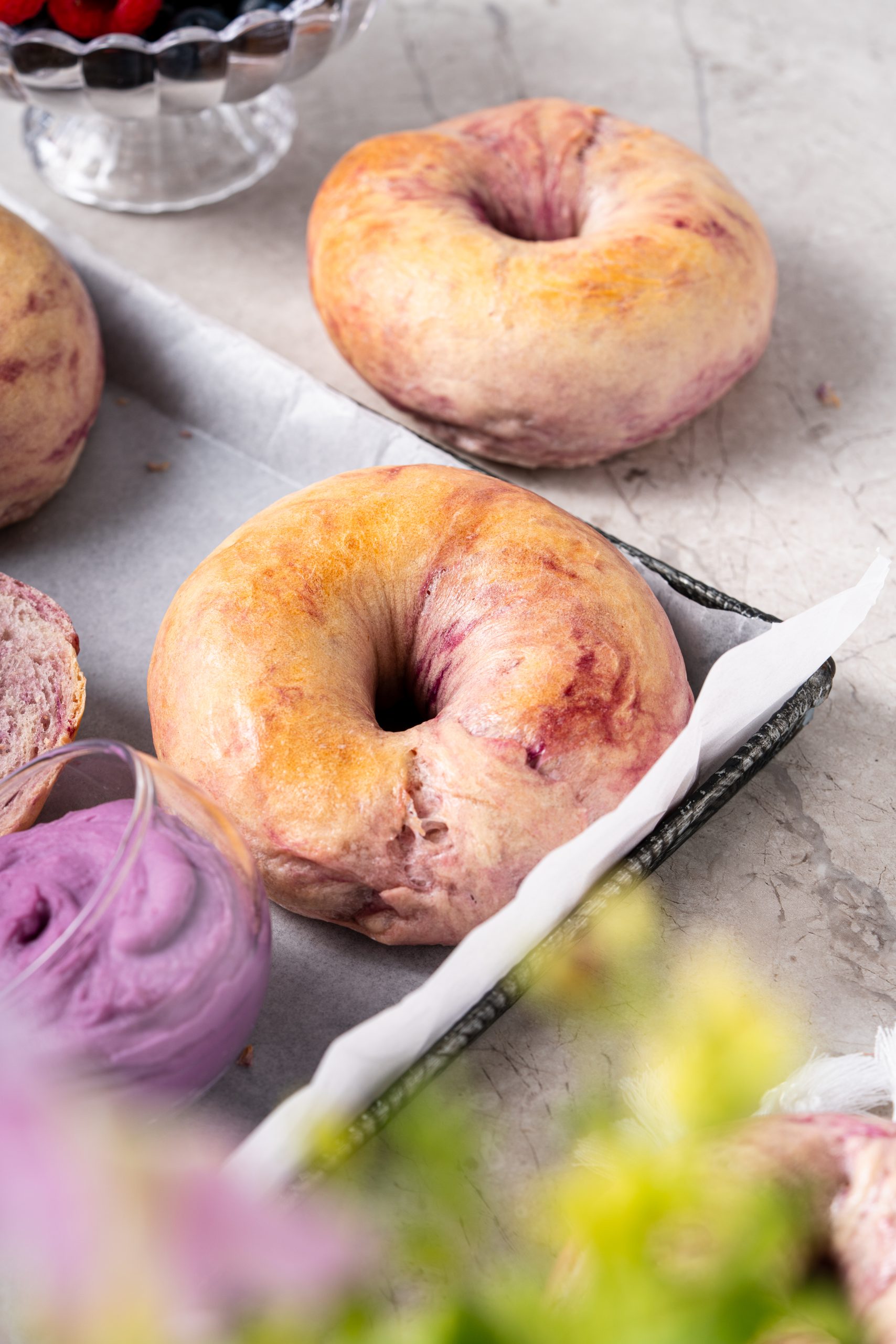 Golden-baked Purple Sweet Potato Bagels resting on a baking tray next to a jar of purple cream and fresh berries.