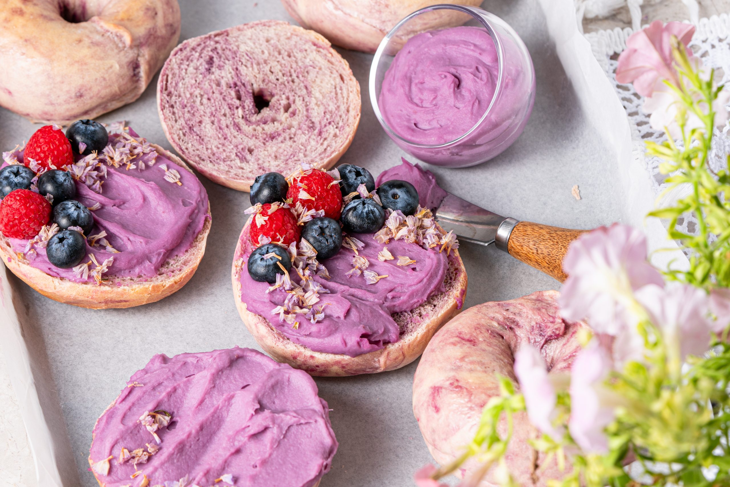 Sliced Purple Sweet Potato Bagels spread with purple cream on a baking tray with fresh berries