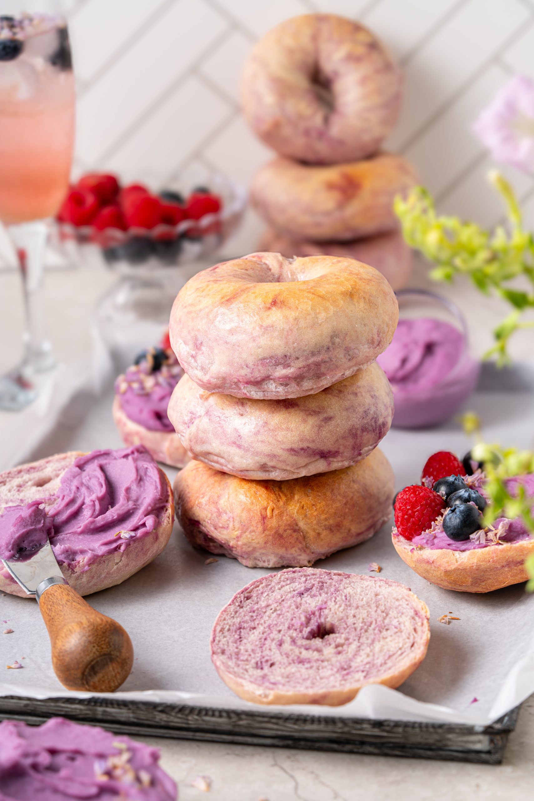 Purple sweet potato bagels stacked on a tray, showing their soft lavender crumb and golden crust, with one sliced open and spread with purple sweet potato cream cheese, topped with fresh raspberries and blueberries.