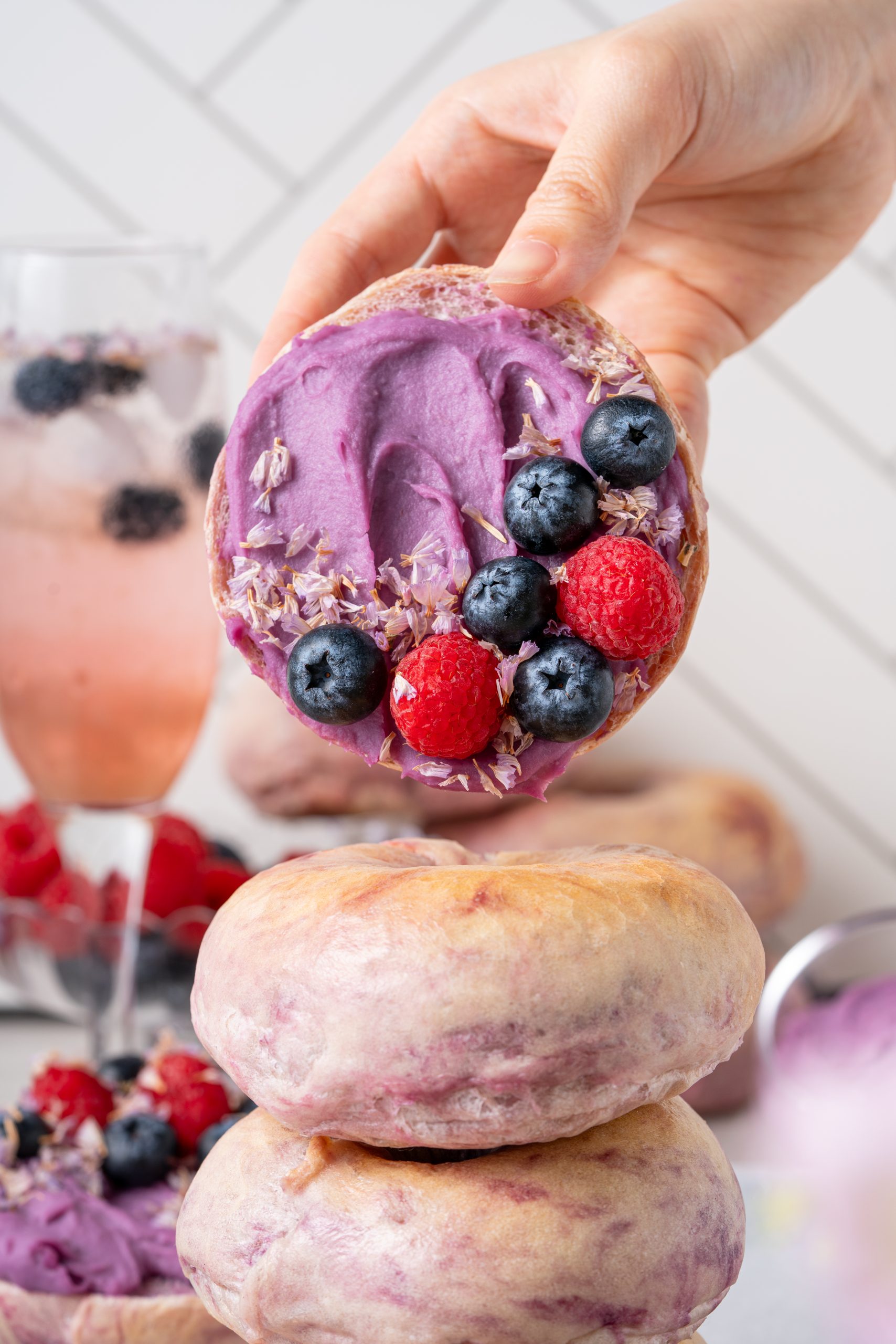 Hand holding a Purple Sweet Potato Bagel topped with vibrant purple spread, blueberries, and raspberries.