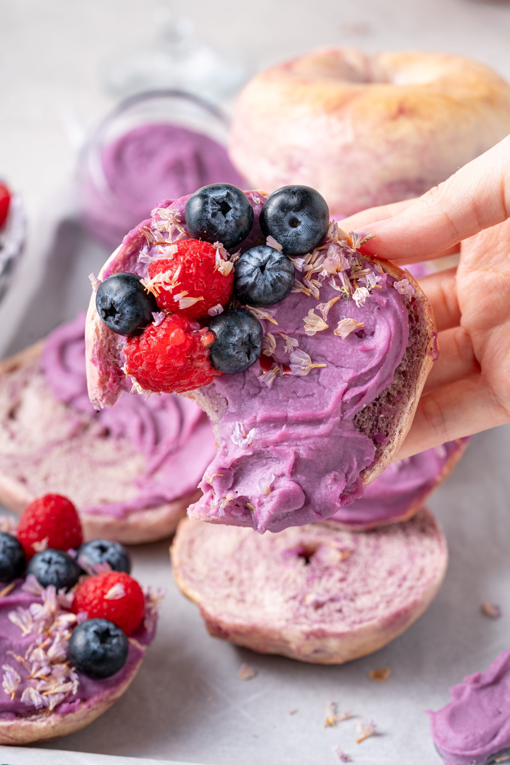 Hand holding a bitten Purple Sweet Potato Bagel with purple sweet potato spread and fresh berries on top.