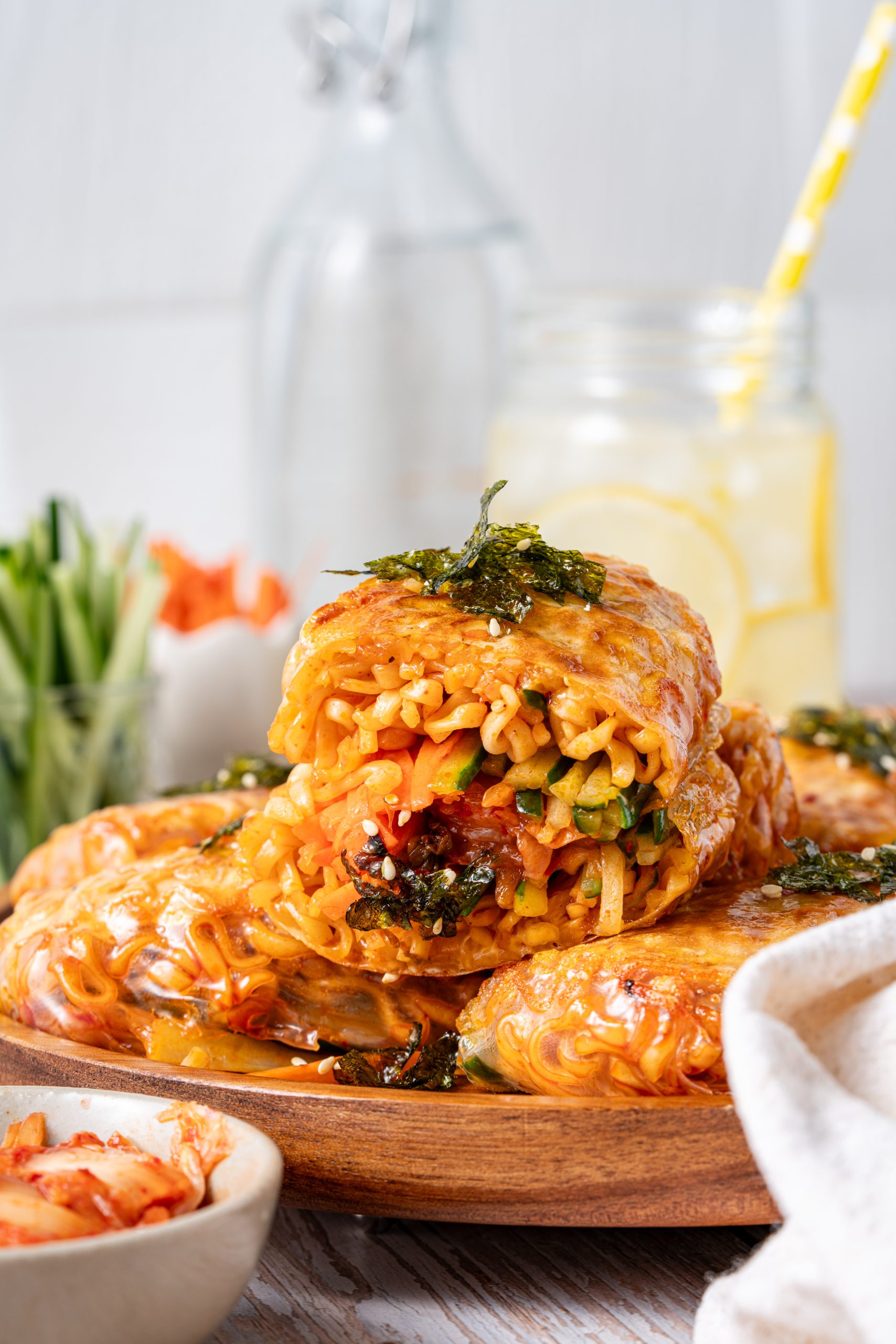A close side view of a ramen spring roll piled on top of others, cut open to show noodles, vegetables, and kimchi spilling slightly from the crispy wrapper.
