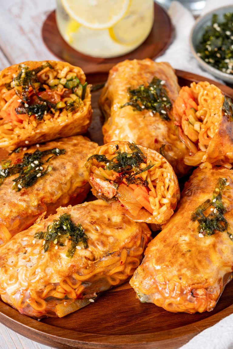 “Close-up of crispy ramen spring rolls filled with noodles, vegetables, and kimchi, topped with toasted seaweed and sesame seeds on a wooden serving plate.”