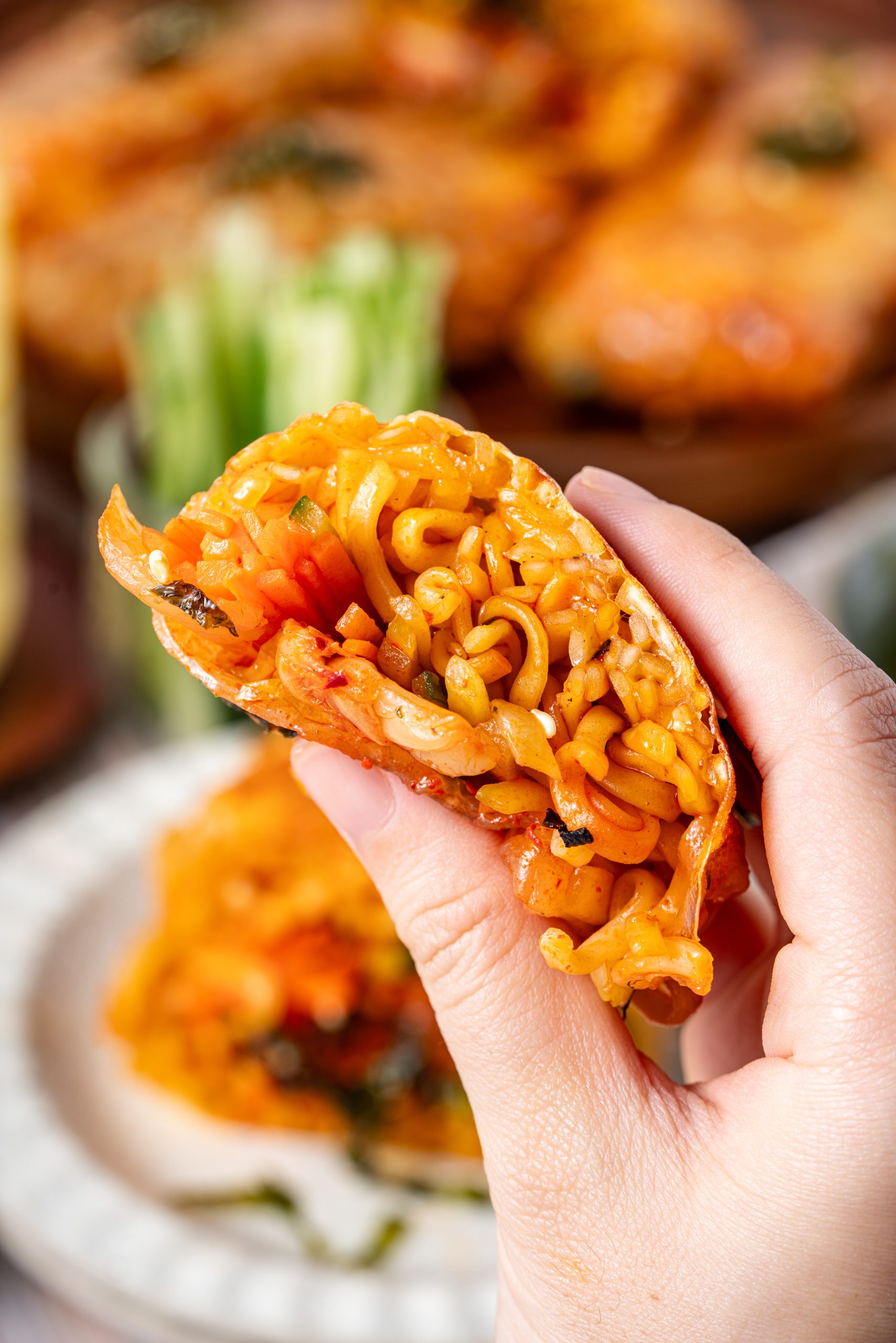 A person holding a half ramen spring roll, displaying the inside filled with saucy cooked noodles, carrots, cucumbers, and kimchi.