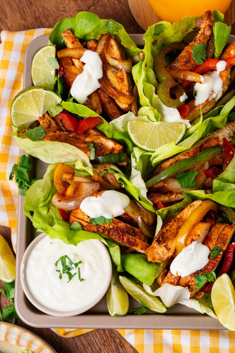 Lettuce wraps filled with chicken fajitas—strips of chicken, bell peppers, and onions—topped with dollops of sour cream and served with lime wedges and a bowl of sour cream on the side.