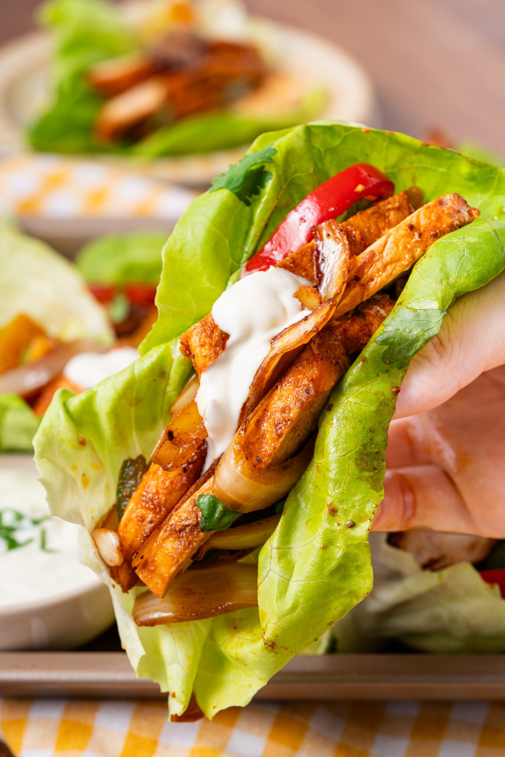 A hand holding a fresh lettuce wrap filled with seasoned chicken strips, sautéed onions and peppers, and a topping of sour cream.