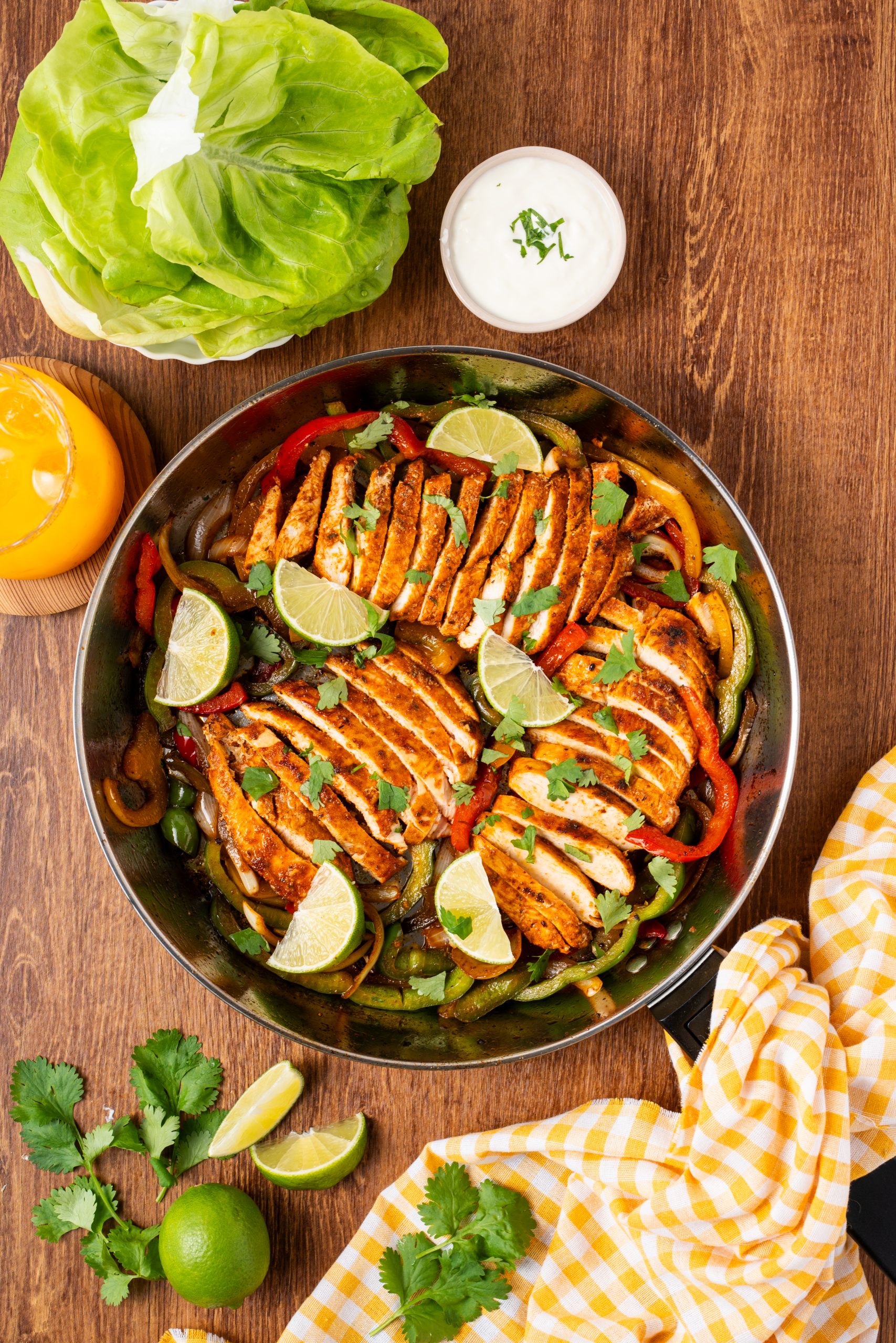 A skillet filled with sliced seasoned chicken, sautéed bell peppers, and onions, topped with lime wedges and fresh cilantro, surrounded by butter lettuce, a bowl of sour cream, and a glass of orange juice on a wooden table.