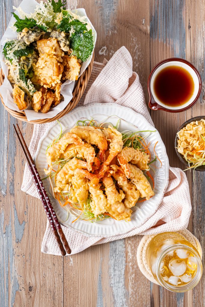 A plate of golden, crispy shrimp tempura arranged on a bed of shredded carrots and green onions, served with a basket of assorted vegetable tempura and a bowl of dipping sauce on a rustic wooden table.