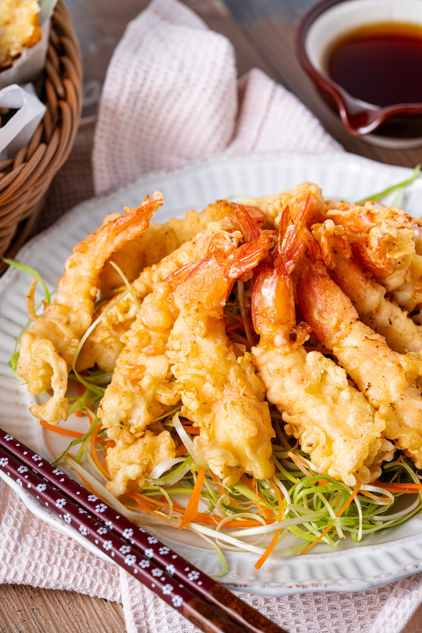 A close-up shot of golden, crispy shrimp tempura arranged on a bed of thinly sliced carrots and green onions, with a bowl of dipping sauce blurred in the background.