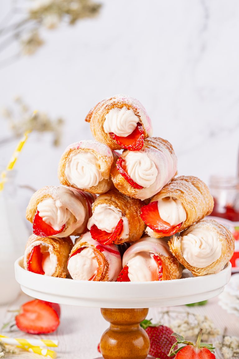 Stack of golden, flaky Cream Horns filled with whipped cream and fresh strawberries, dusted with powdered sugar and drizzled with pink icing on a cake stand.