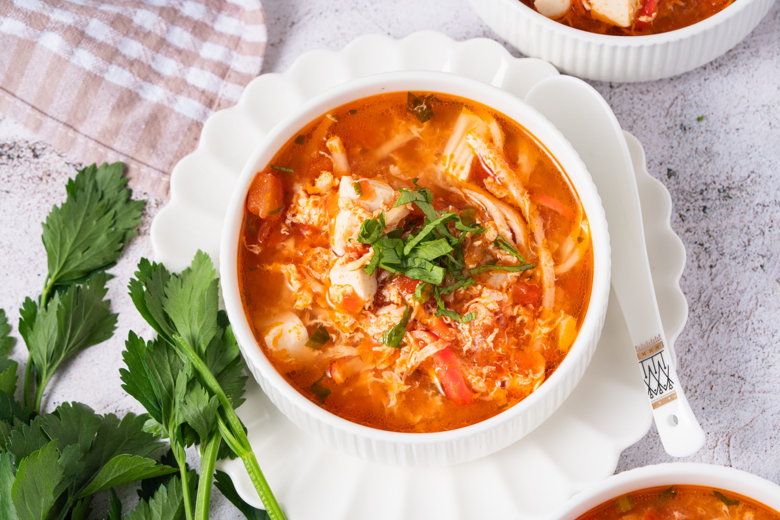 Overhead shot of a single bowl of Tomato Egg Drop Soup showcasing the bright red broth with soft egg strands, diced tofu, and tomatoes, garnished with fresh herbs beside whole tomatoes and eggs.