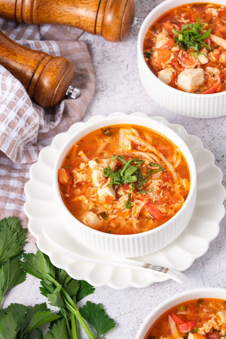 A close-up of Tomato Egg Drop Soup served in a white ribbed bowl, filled with silky egg ribbons, tofu cubes, and tomato chunks, topped with chopped herbs on a rustic white surface.