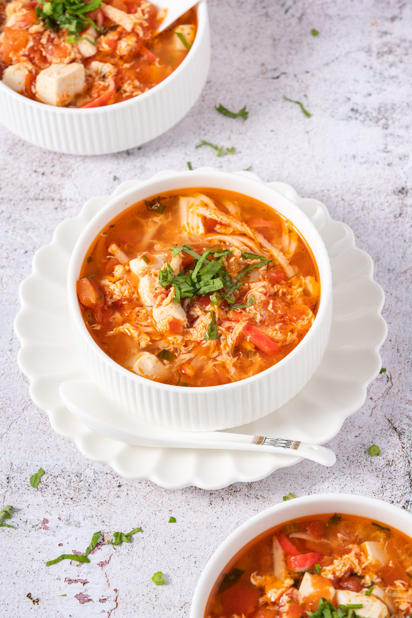 A close-up of Tomato Egg Drop Soup served in a white ribbed bowl, filled with silky egg ribbons, tofu cubes, and tomato chunks, topped with chopped herbs on a rustic white surface.