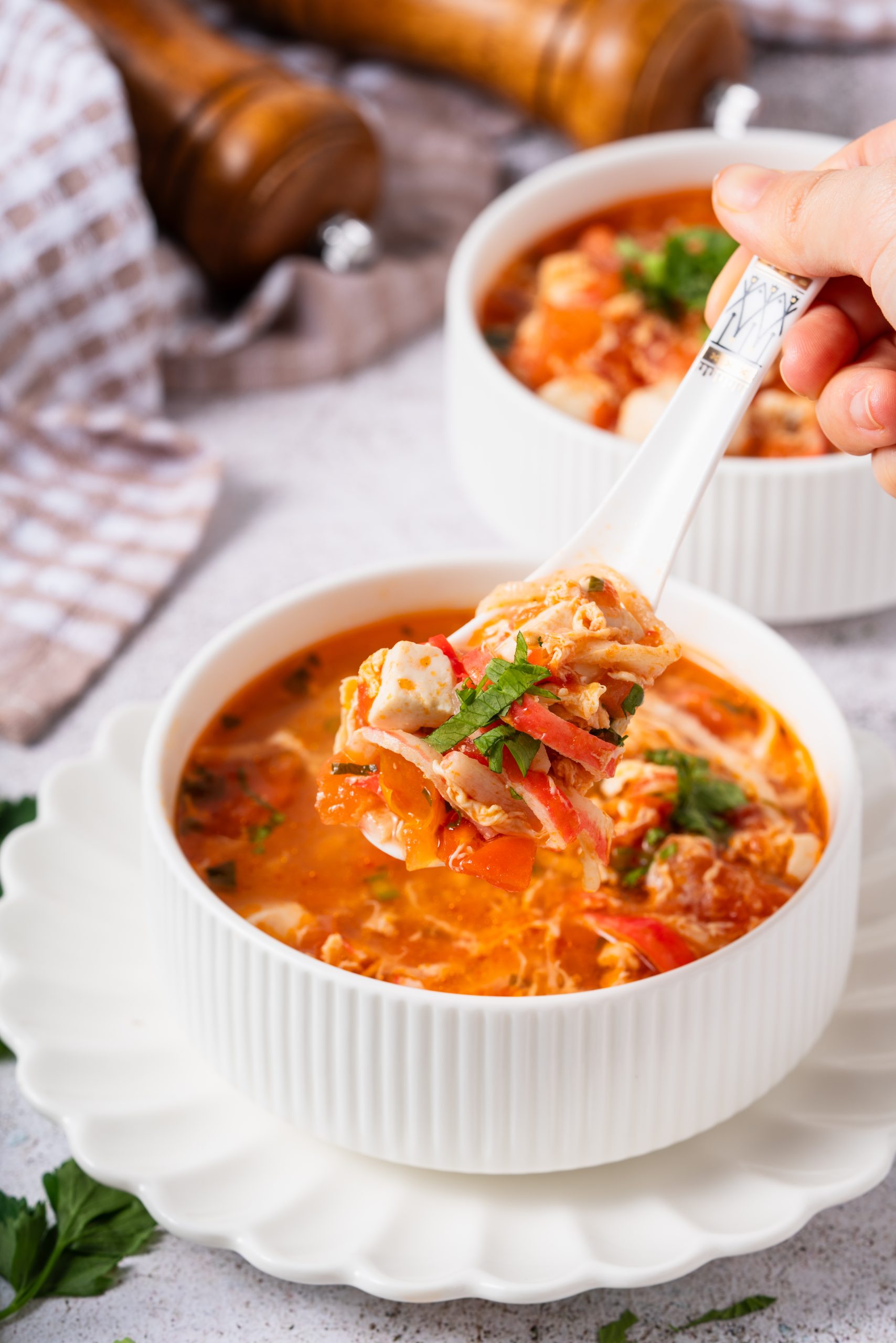 A hand holding a spoonful of hearty Tomato Egg Drop Soup filled with tofu, shredded egg, and tomatoes, above a bowl placed on a scalloped white plate.