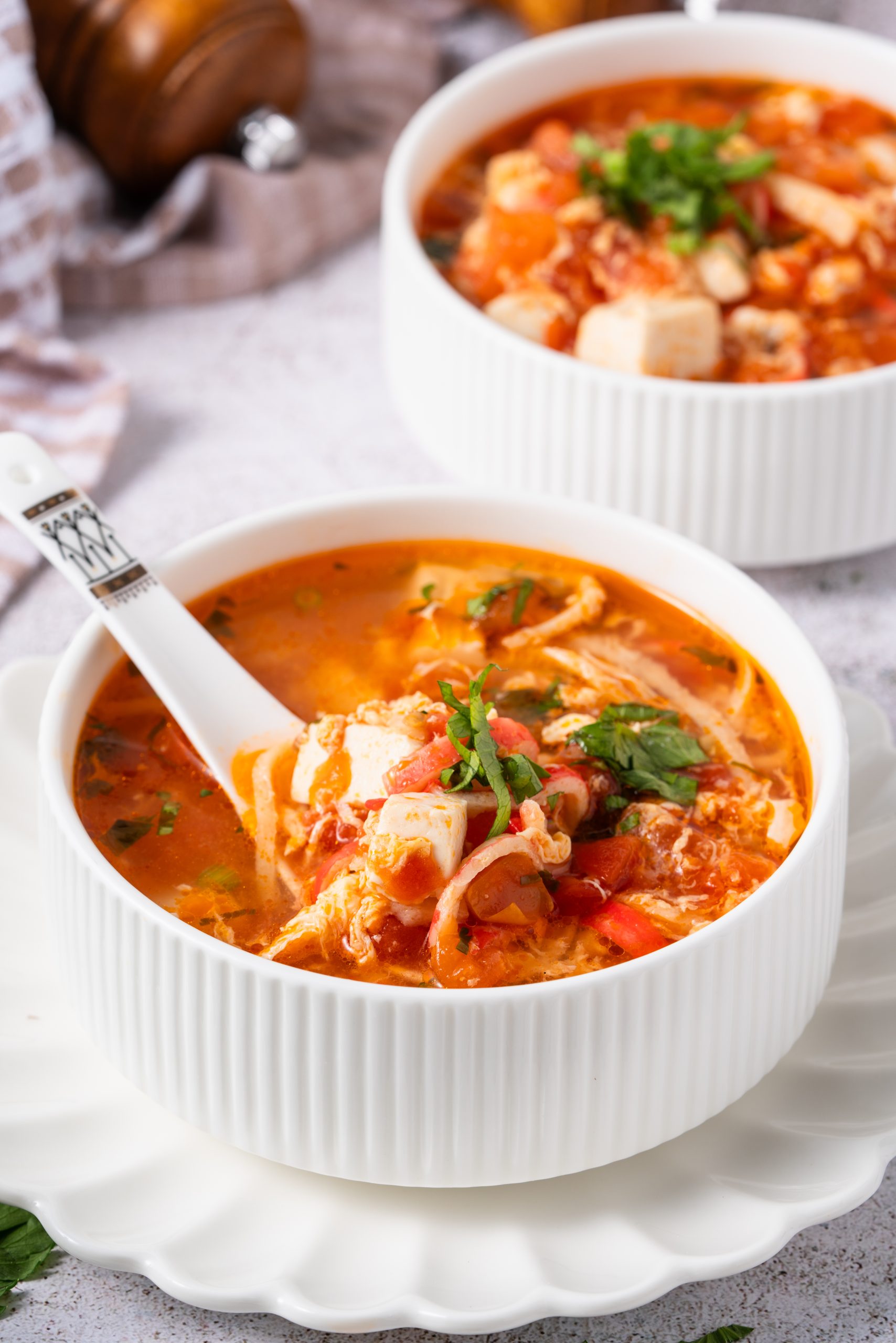 Close-up of a white bowl filled with Tomato Egg Drop Soup, showing silky egg ribbons, diced tofu, and tomato chunks in a rich red broth, garnished with chopped herbs and served with a white ceramic spoon.