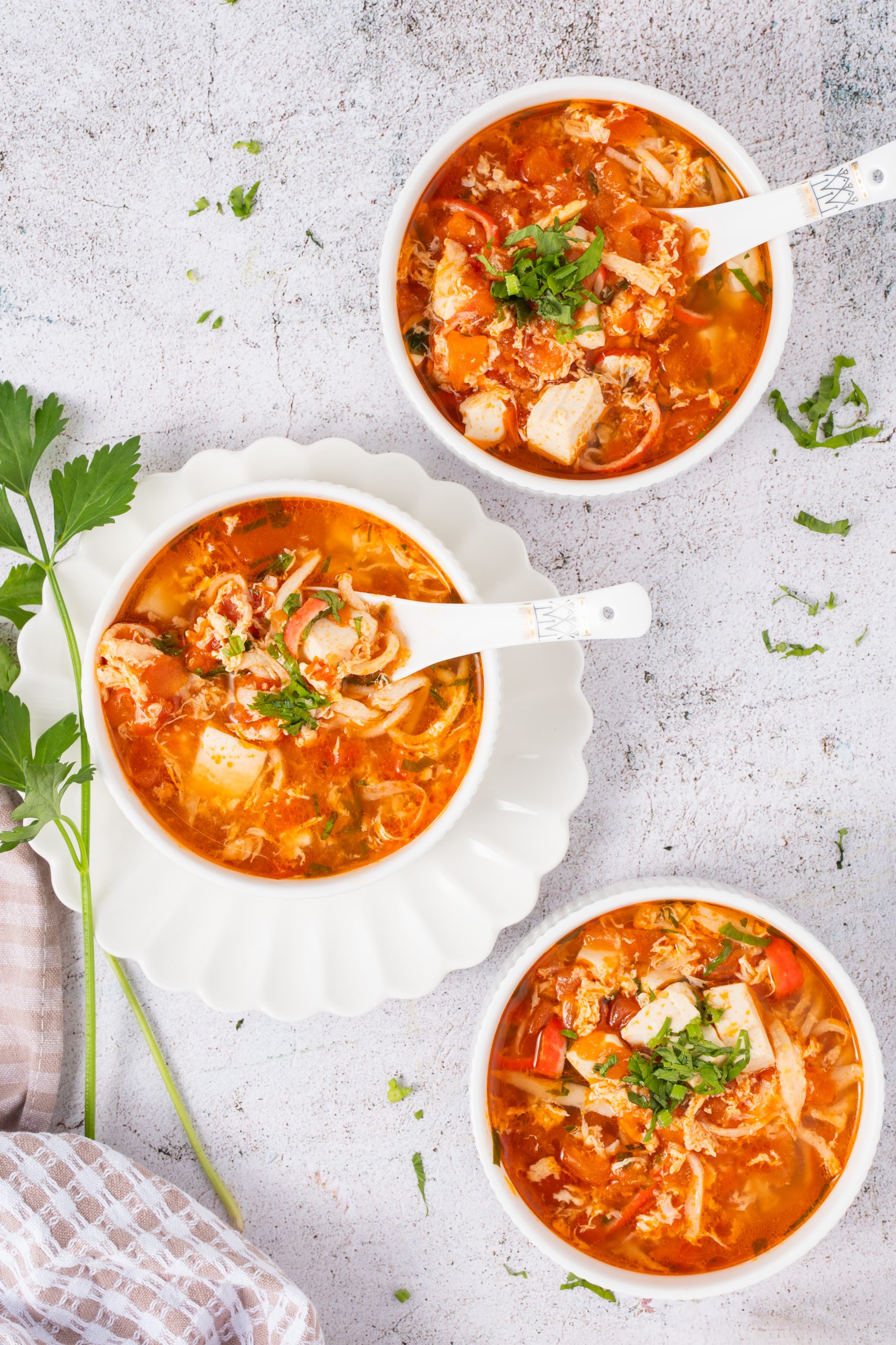 Flat lay of three bowls of Tomato Egg Drop Soup, each filled with tofu, egg, and tomato in a rich red broth, garnished with herbs and served with white ceramic spoons.