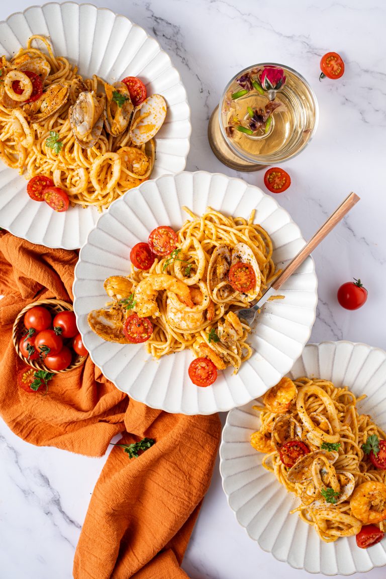 Overhead view of three plates of tom yum seafood pasta arranged around an orange cloth, with cherry tomatoes scattered around.