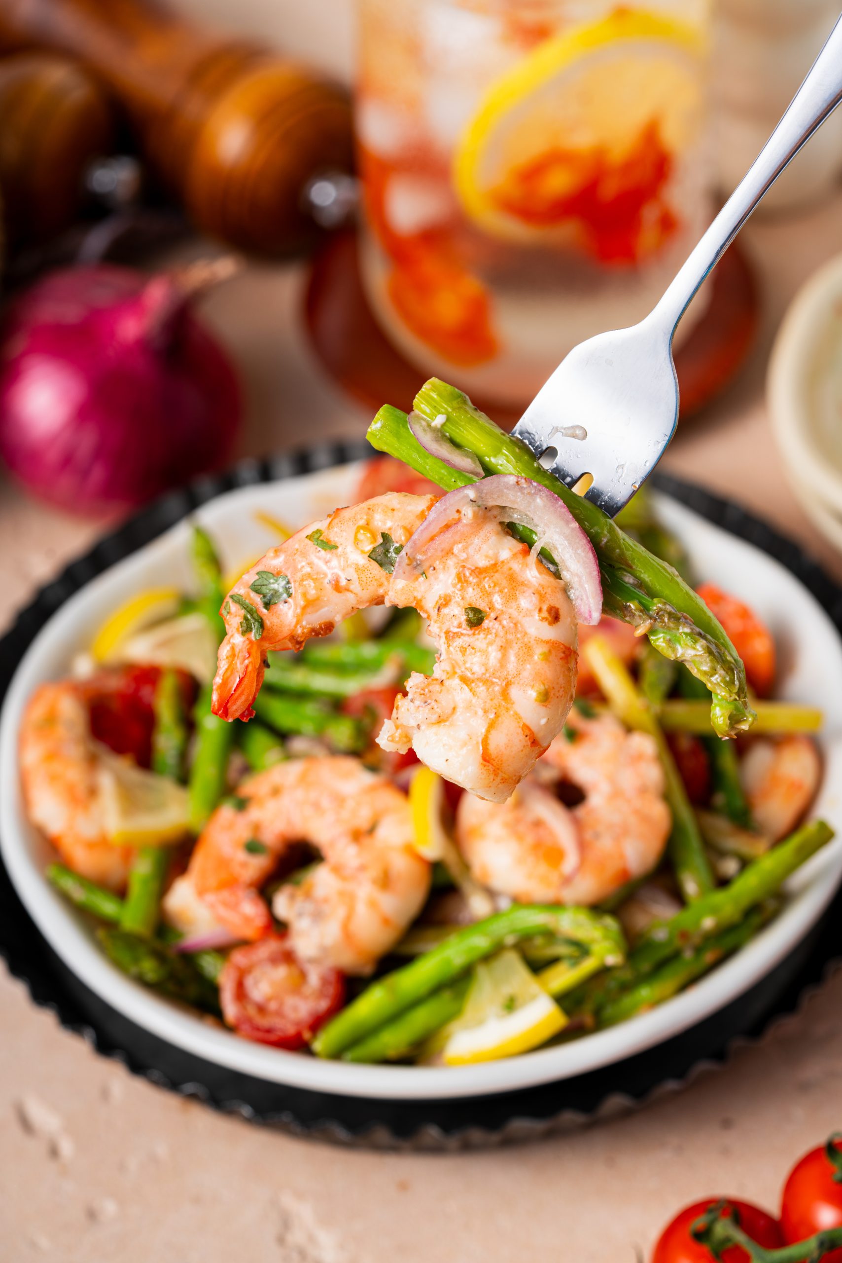 A fork lifting shrimp and asparagus from a bowl of warm salad with tomatoes and onions, showing the bright and fresh textures.