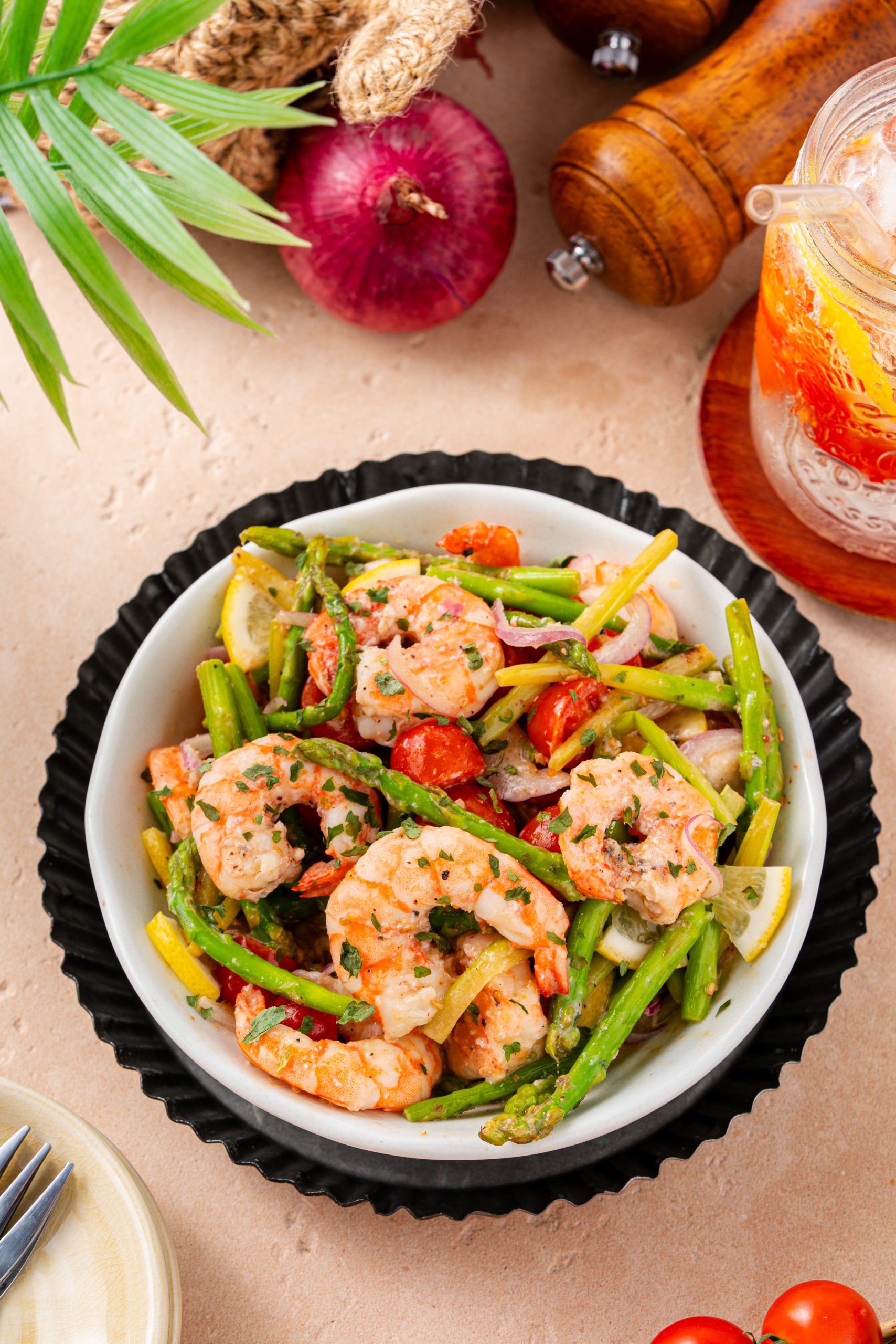 Overhead view of a bowl of warm asparagus and shrimp salad with cherry tomatoes, red onions, and lemon slices, served next to a cold drink.