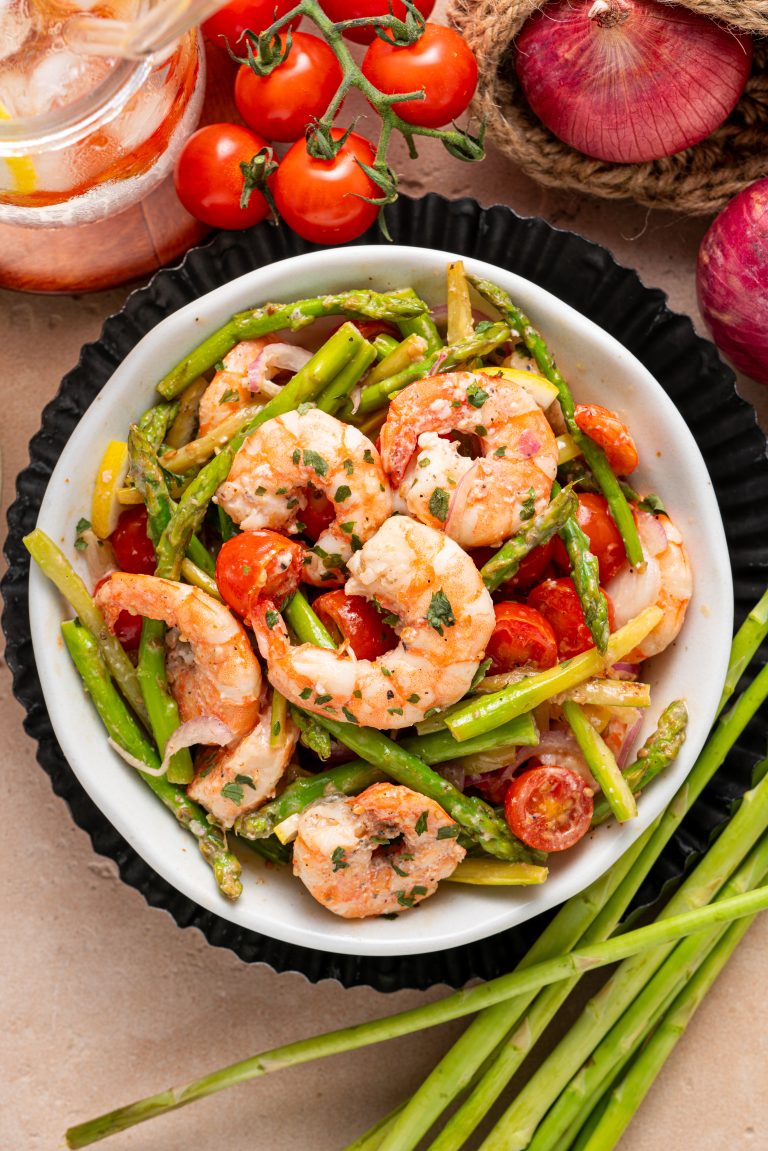 A flat lay of warm asparagus and shrimp salad with tomatoes, onions, and lemon wedges, surrounded by fresh ingredients