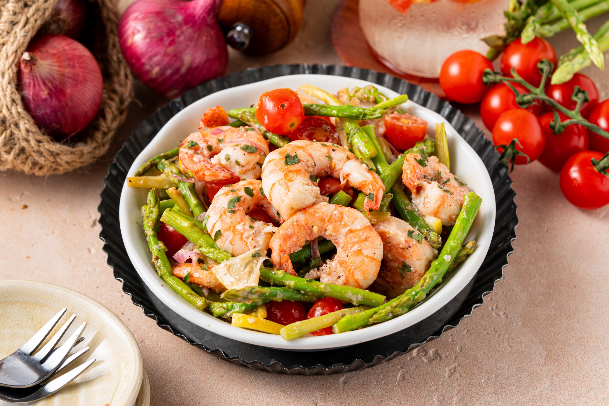 Side angle of warm asparagus and shrimp salad with tomatoes and lemon slices, placed on a rustic table with fresh vegetables in the background.