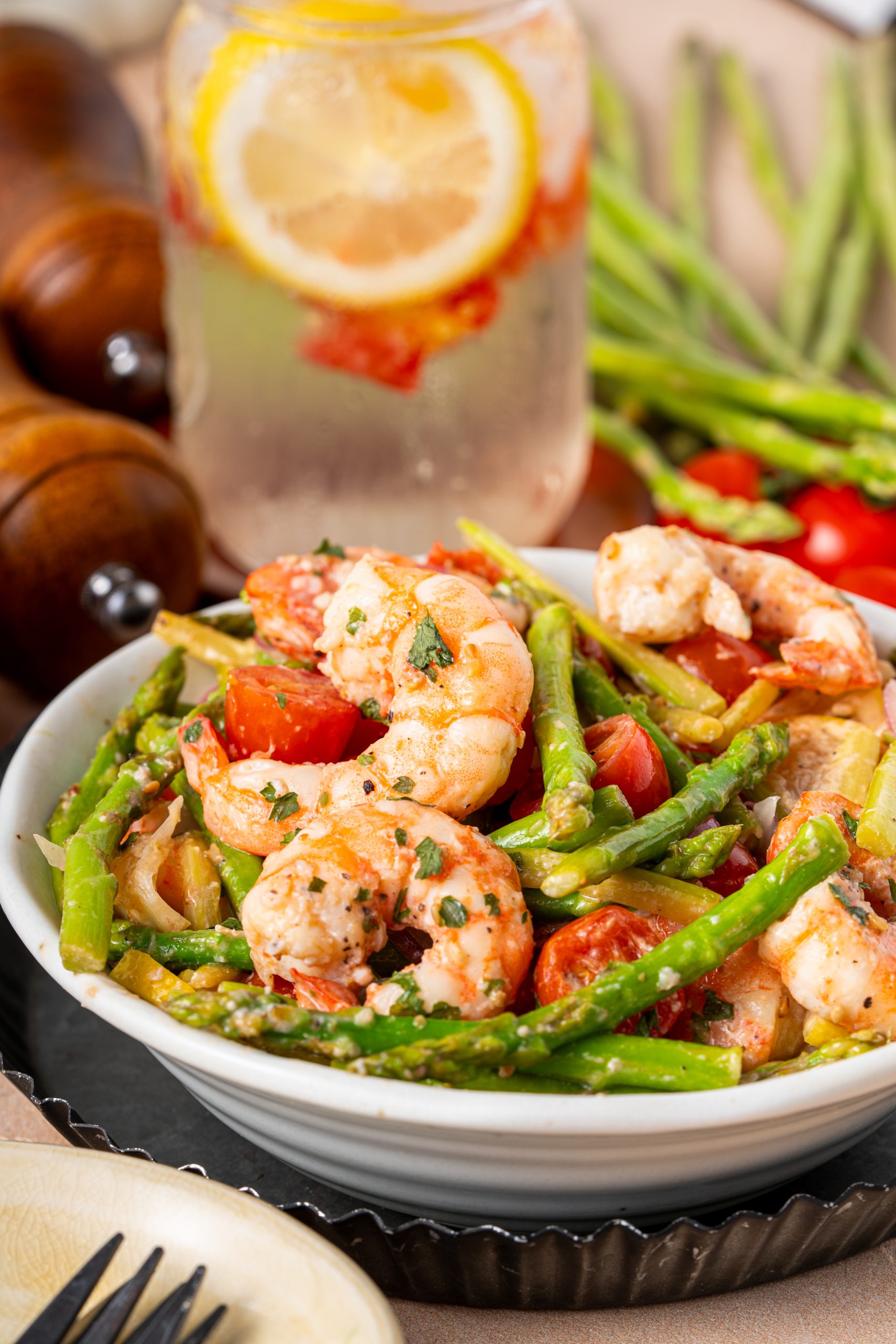 Warm asparagus and shrimp salad with tomatoes, onions, and lemon wedges, surrounded by fresh ingredients and rustic decor.
