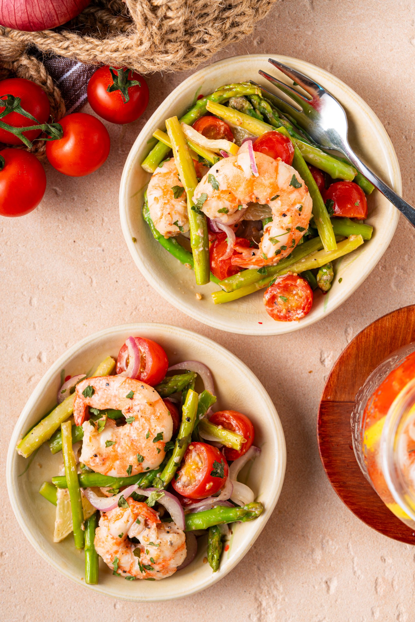Two small plates of warm asparagus and shrimp salad with tomatoes and red onions, styled on a beige tabletop with fresh produce.