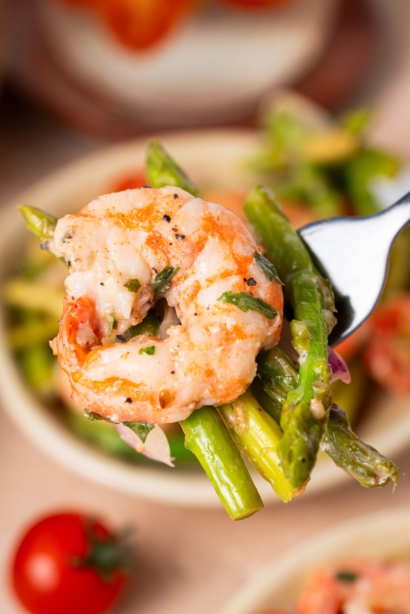A fork lifting shrimp and asparagus from a bowl of warm salad with tomatoes and onions, showing the bright and fresh textures.