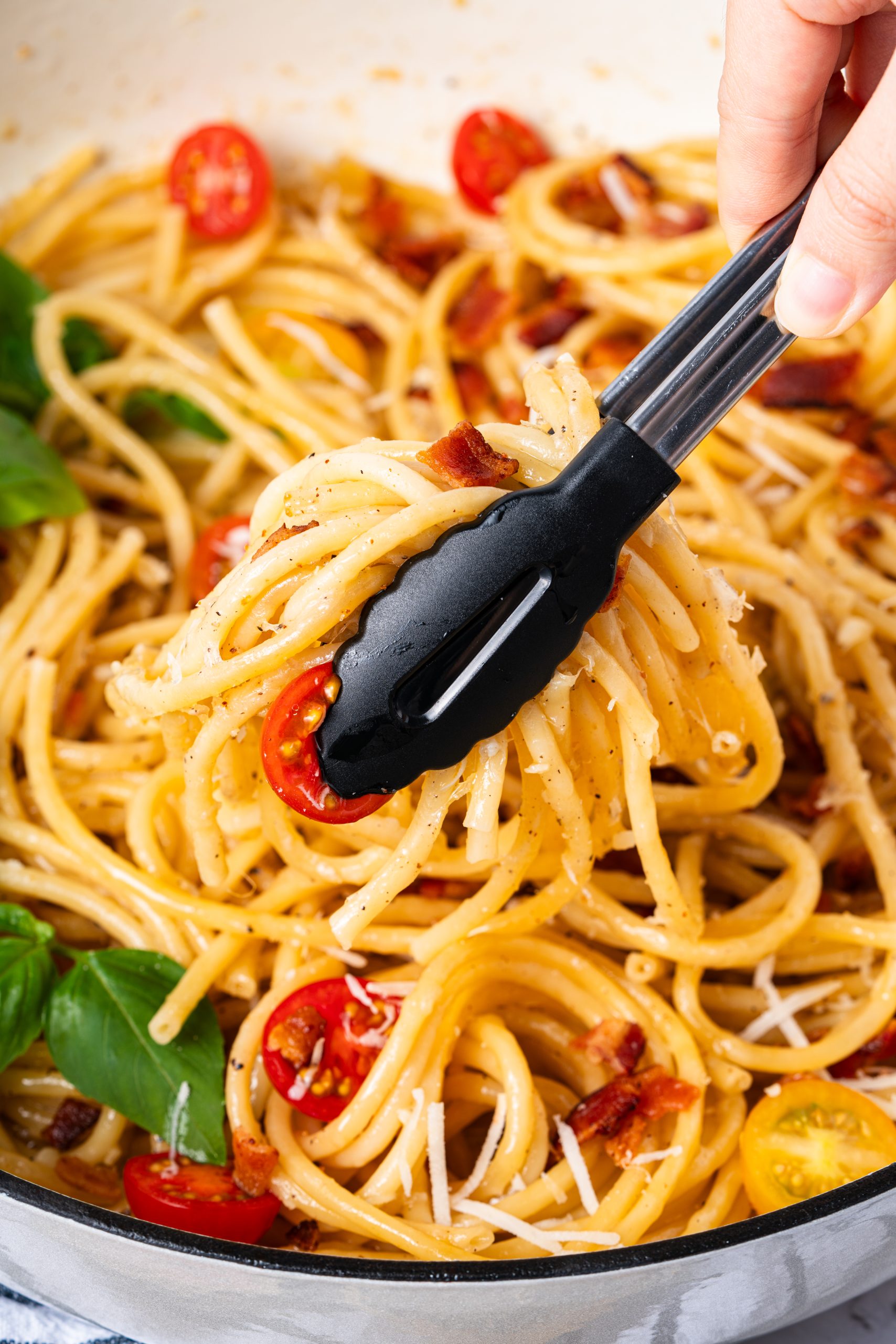 Close-up of tongs lifting Cacio e Pepe spaghetti, showing glossy cheese sauce, black pepper, and tomato slices.