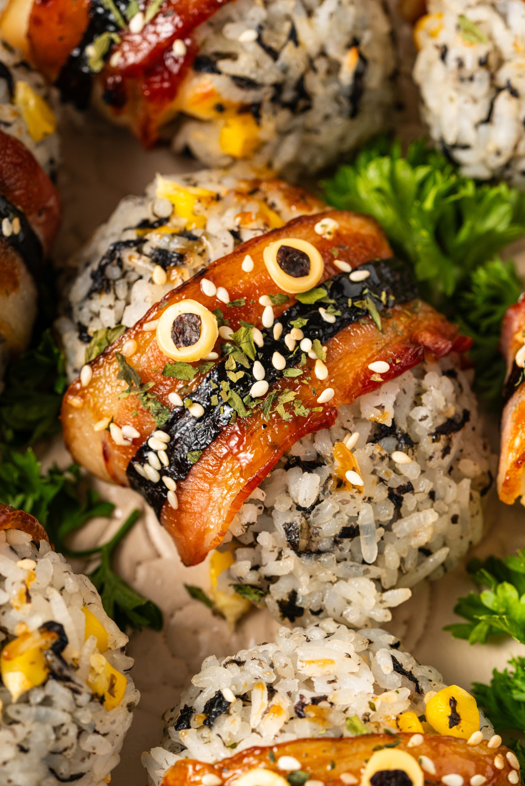 Close-up of bacon-wrapped rice balls topped with sesame seeds and nori, featuring crispy glazed bacon over seasoned rice with corn, garnished with fresh parsley.