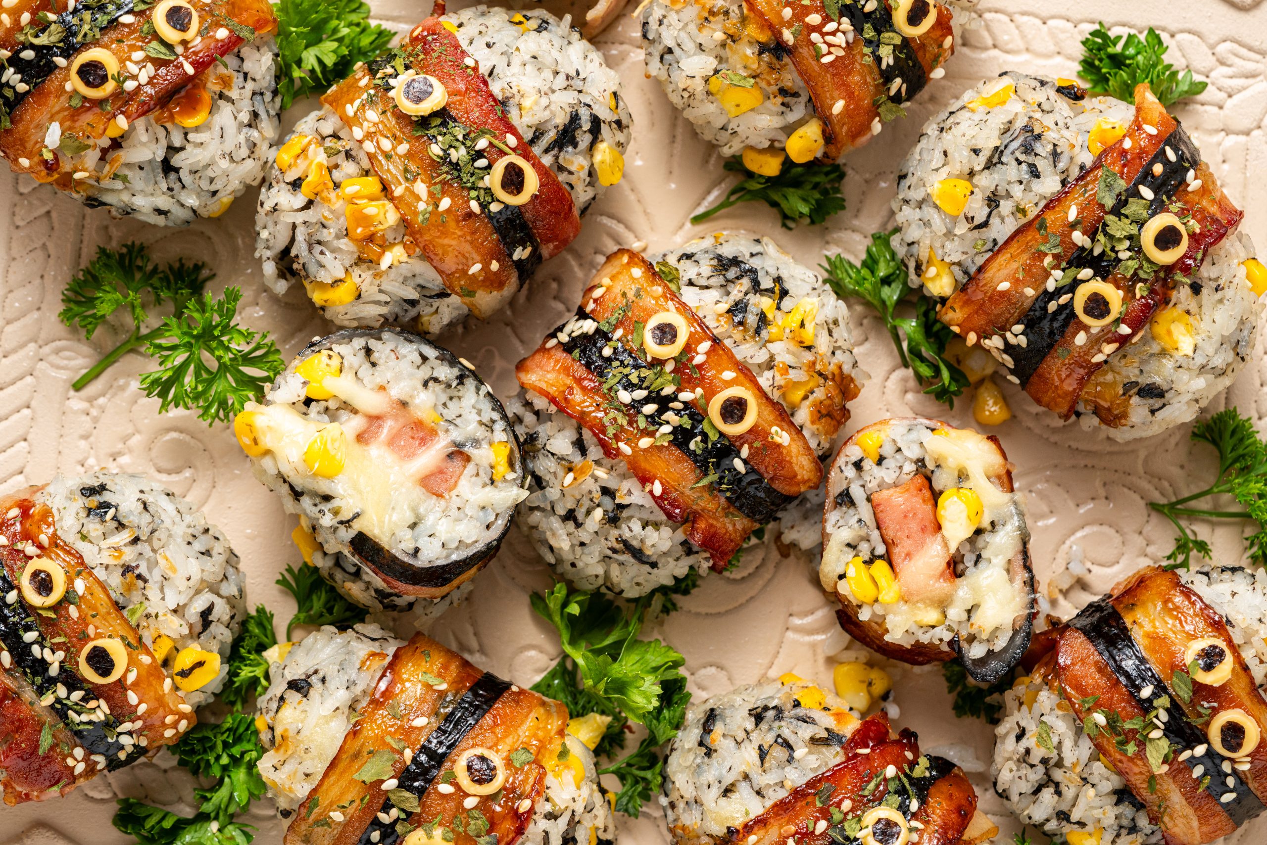 Overhead view of bacon-wrapped rice balls topped with sesame seeds and herbs on a ceramic platter
