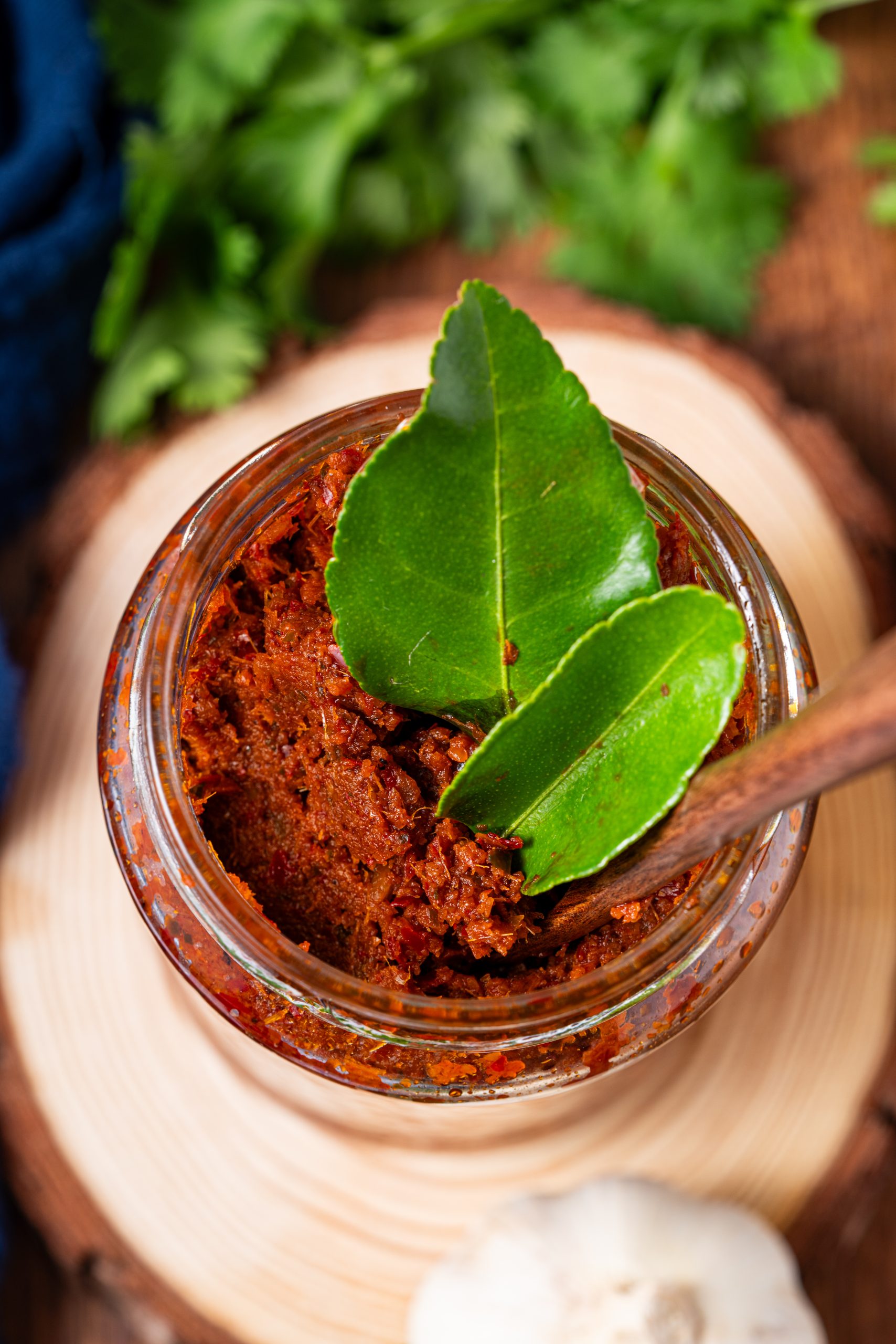 Panang curry paste in jar with lime leaves, garlic, dried chilies, and peanuts on wooden table.