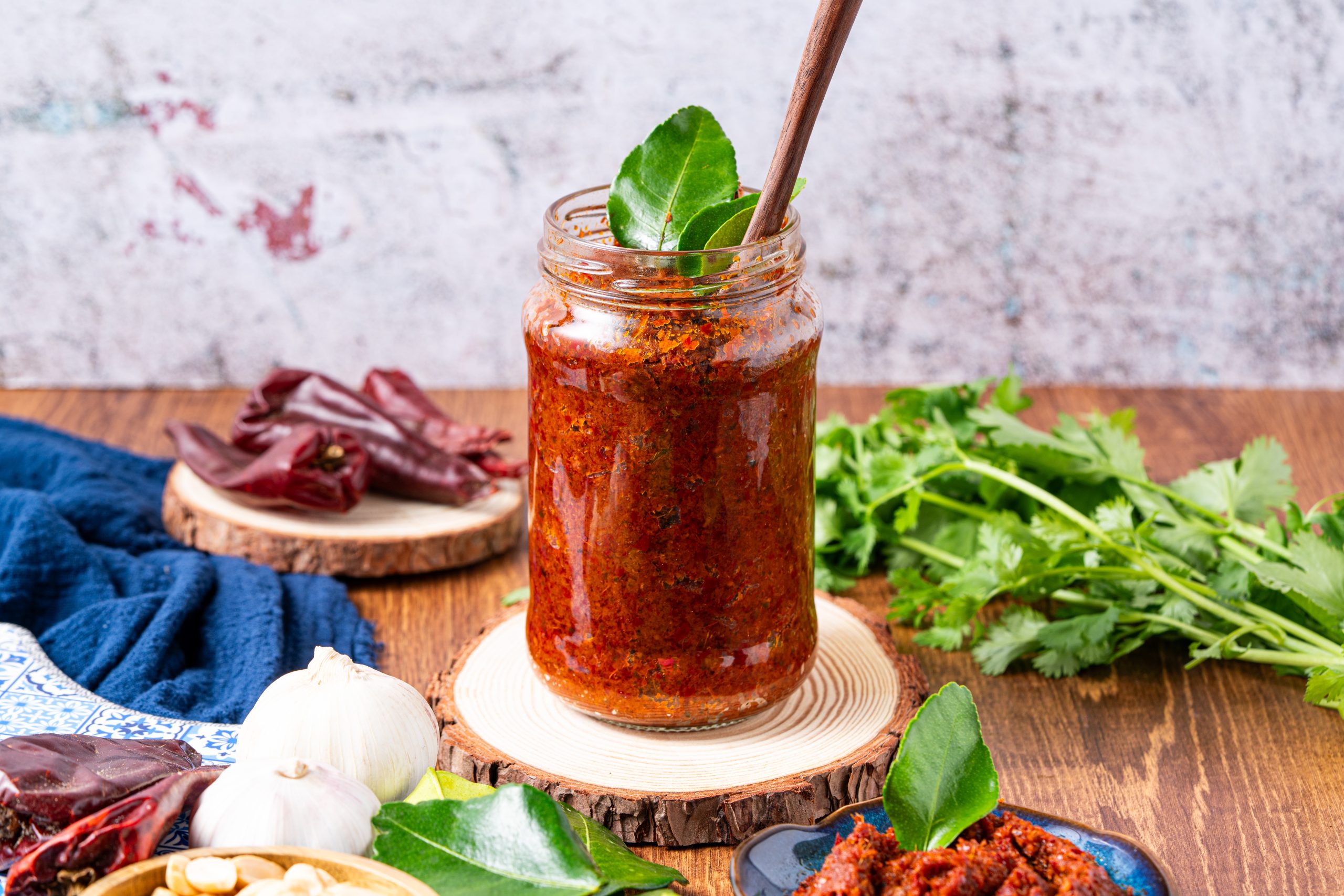 Jar of Thai Panang curry paste with wooden spoon, surrounded by fresh herbs and spices.
