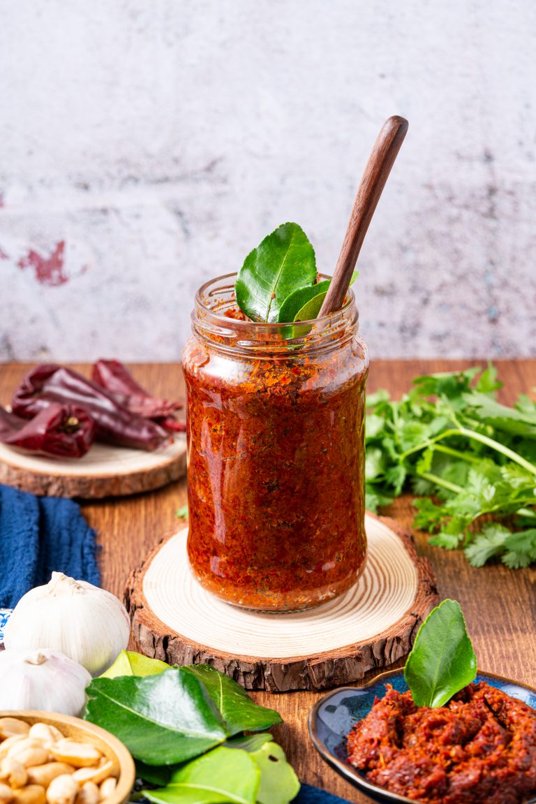 Panang curry paste in glass jar with kaffir lime leaves, dried red chilies, and garlic.