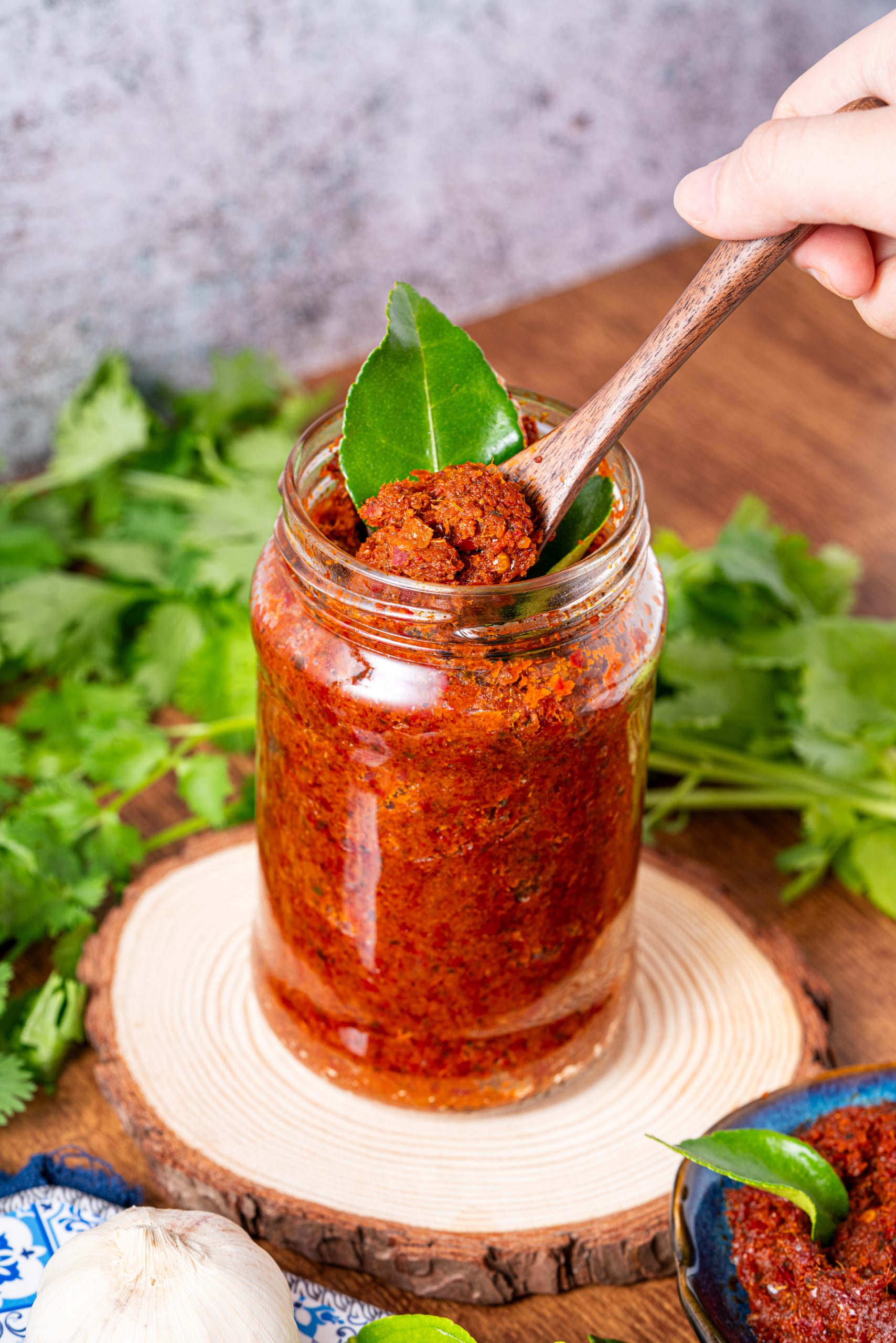 Image 2:
Homemade Panang curry paste in a glass jar with wooden spoon and kaffir lime leaves, surrounded by garlic and fresh cilantro – rich, spicy Thai curry base.