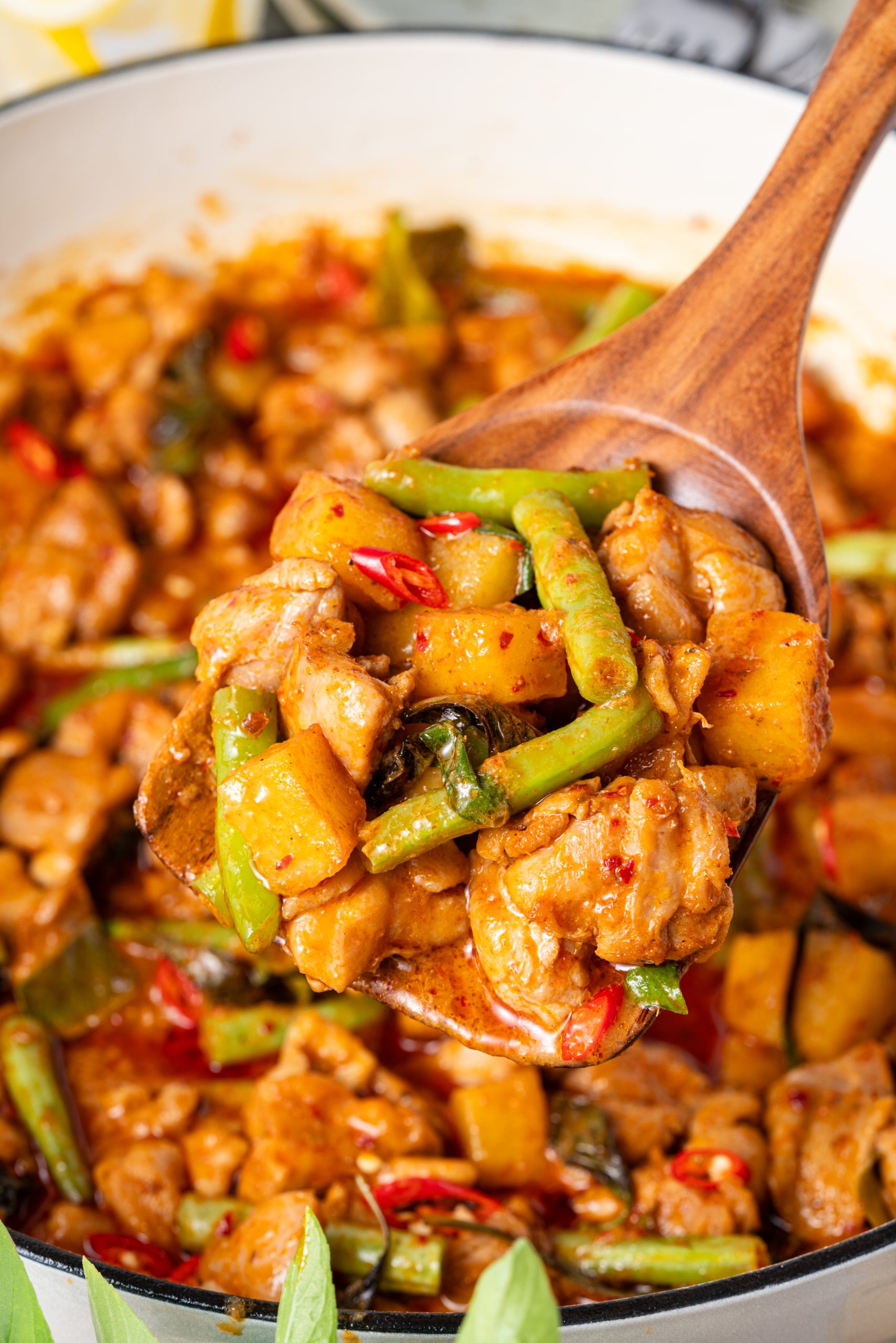 Wooden spoon lifting chicken Panang curry, showing chunks of chicken, pineapple, long beans, and red chilies coated in thick, aromatic Thai curry sauce.